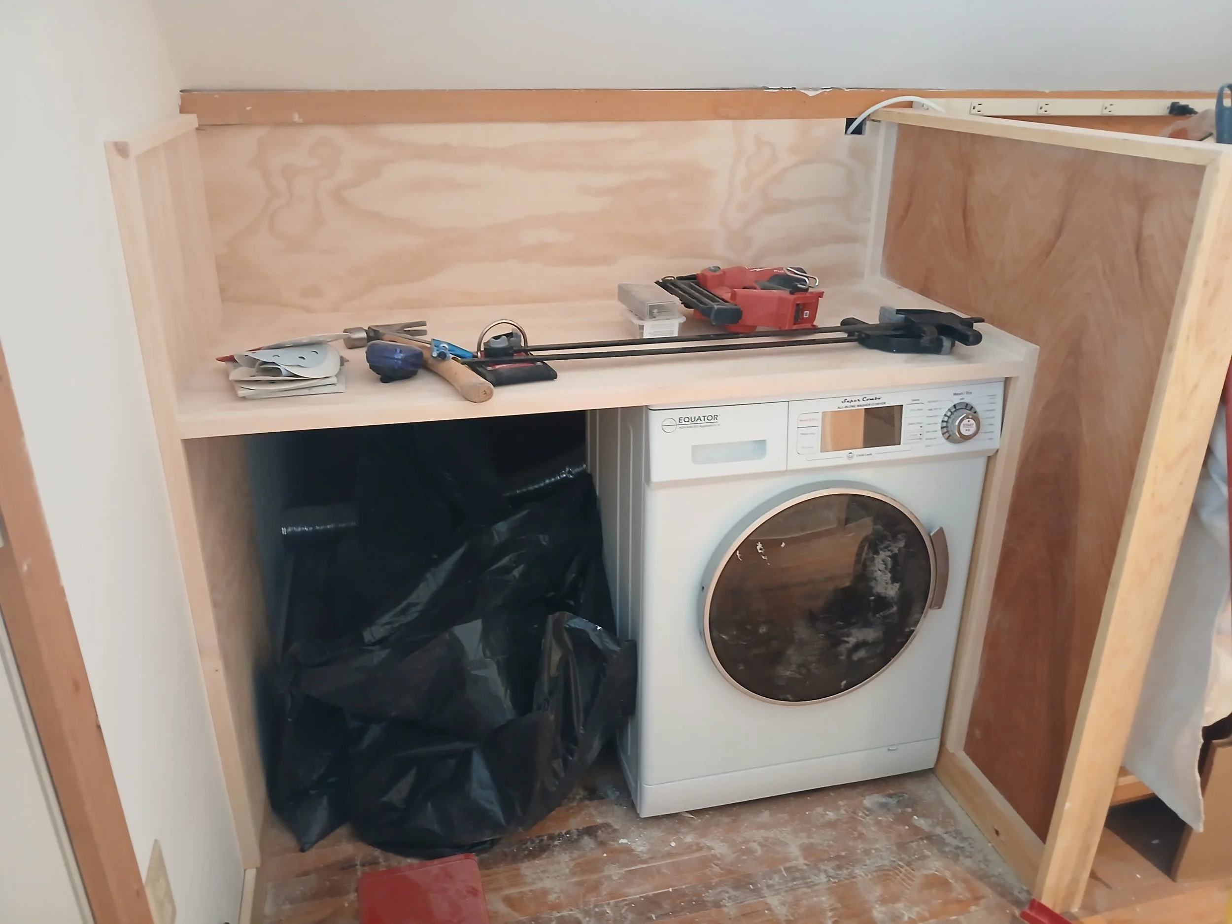 Laundry room with a washing machine installed inside a wooden frame under construction, tools and materials on top of the frame, and a large black trash bag on the floor.