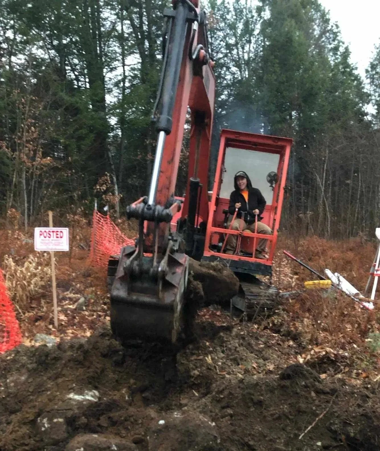 A person operating an orange excavator on a hillside with trees in the background, a 'POSTED' sign on the left, and orange safety fencing.