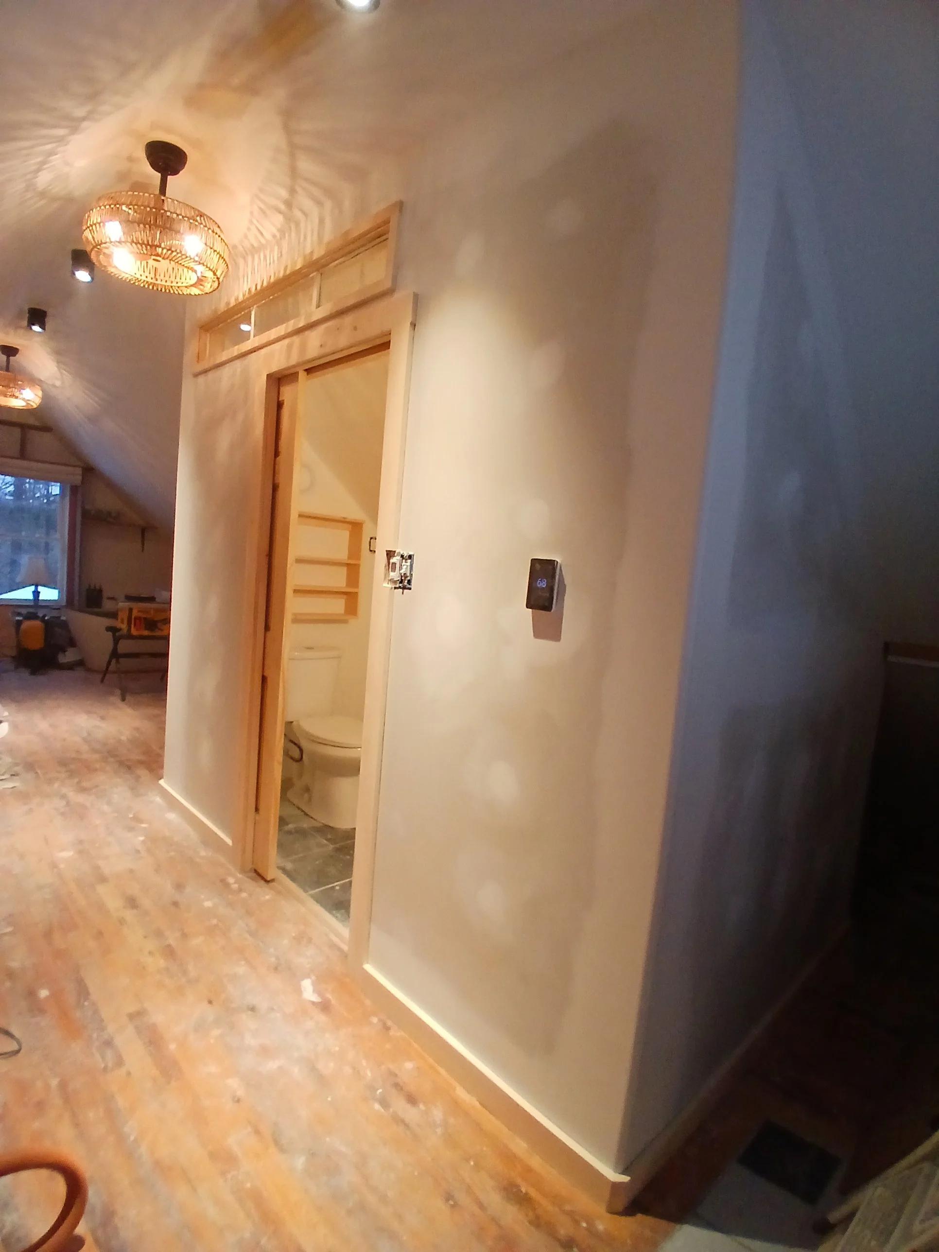 Interior of a house with a hallway leading to a small bathroom, partially visible. The bathroom has a toilet, and the wall framing around the bathroom doorway is unfinished wood. The floor is wooden with some construction dust, and there are two ceil