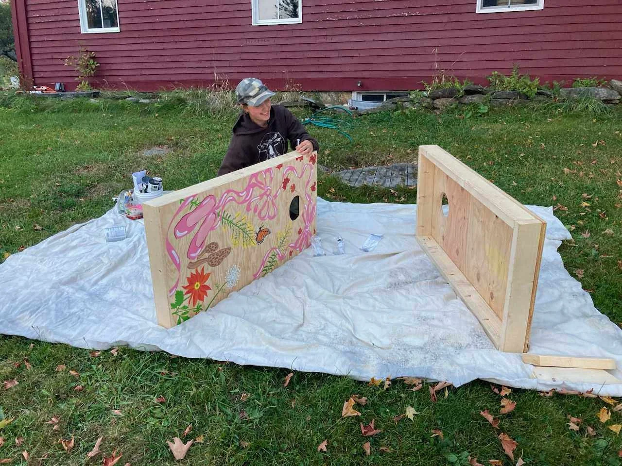 A person painting a decorated wooden panel outdoors on a white sheet, with another plain wooden panel nearby, in front of a red house with white windows.