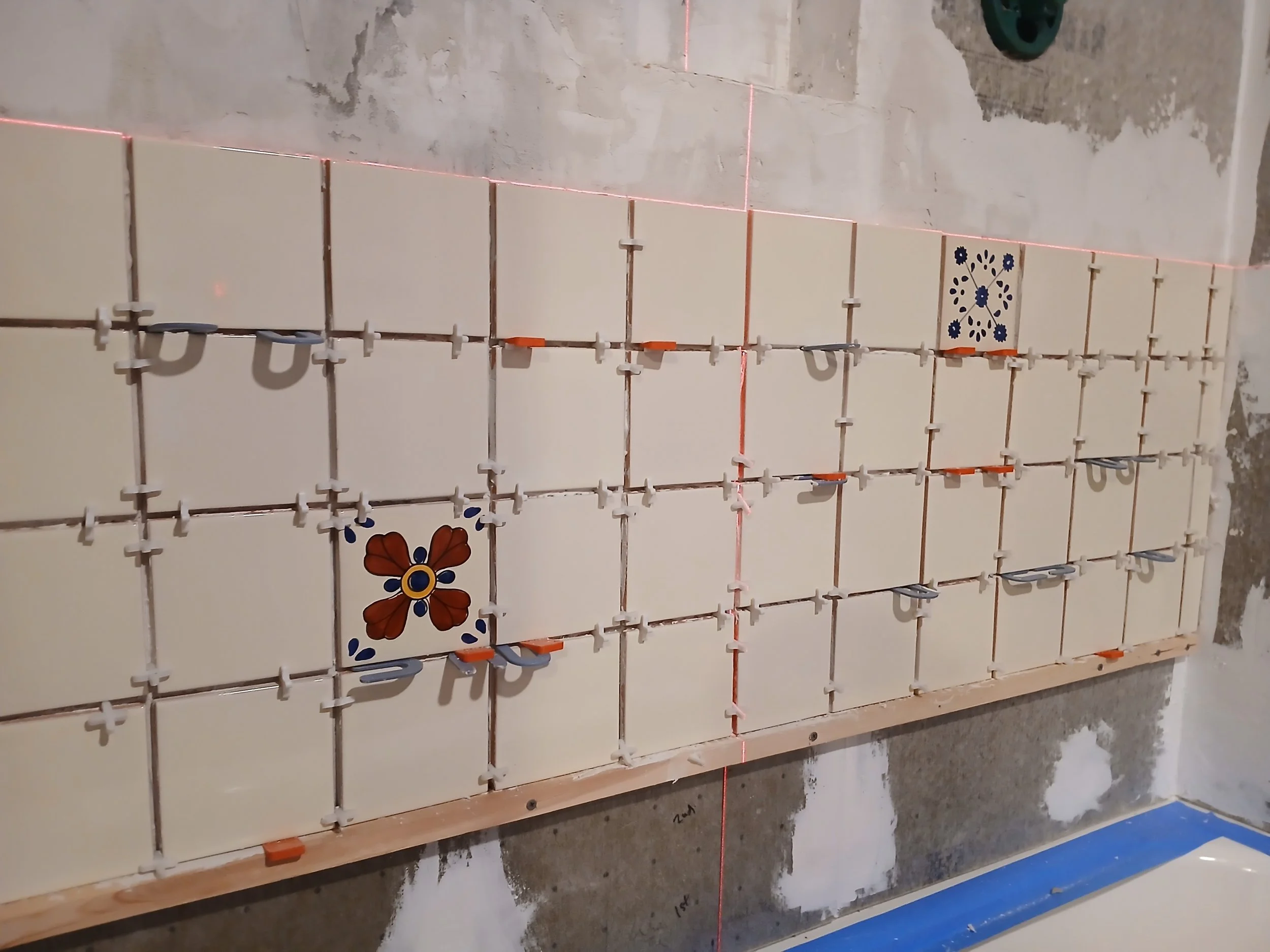 Tile installation in progress on a wall, with partially attached decorative tiles among plain beige tiles, some tiles have floral patterns. The wall has exposed concrete and unfinished areas, with reddish laser guide lines and orange tile spacers.