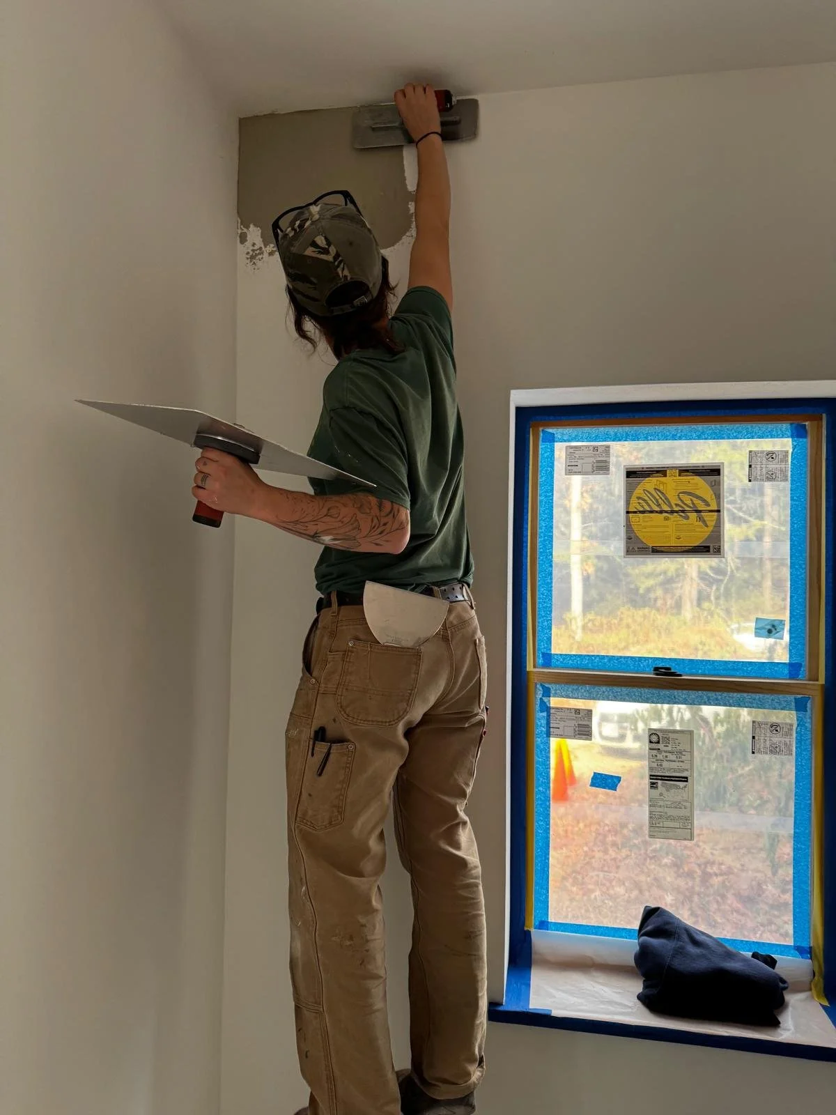 Person installing drywall near a ceiling with a finishing tool, standing on the floor beside a window covered with blue painter's tape.