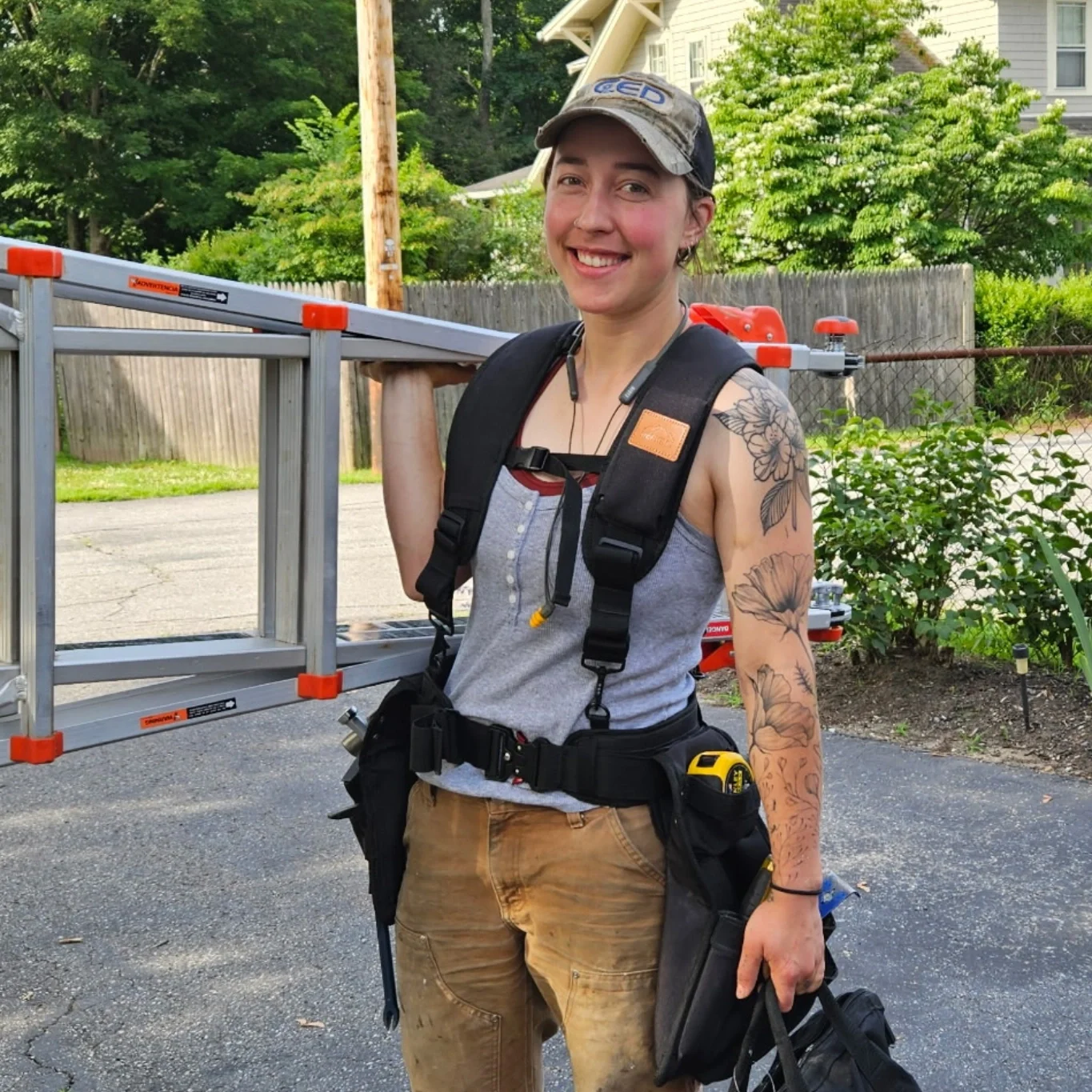 A woman dressed as a construction worker holding a ladder, wearing a gray tank top, brown pants, a tool belt, and a cap, standing outdoors on a paved surface with greenery and a house in the background.