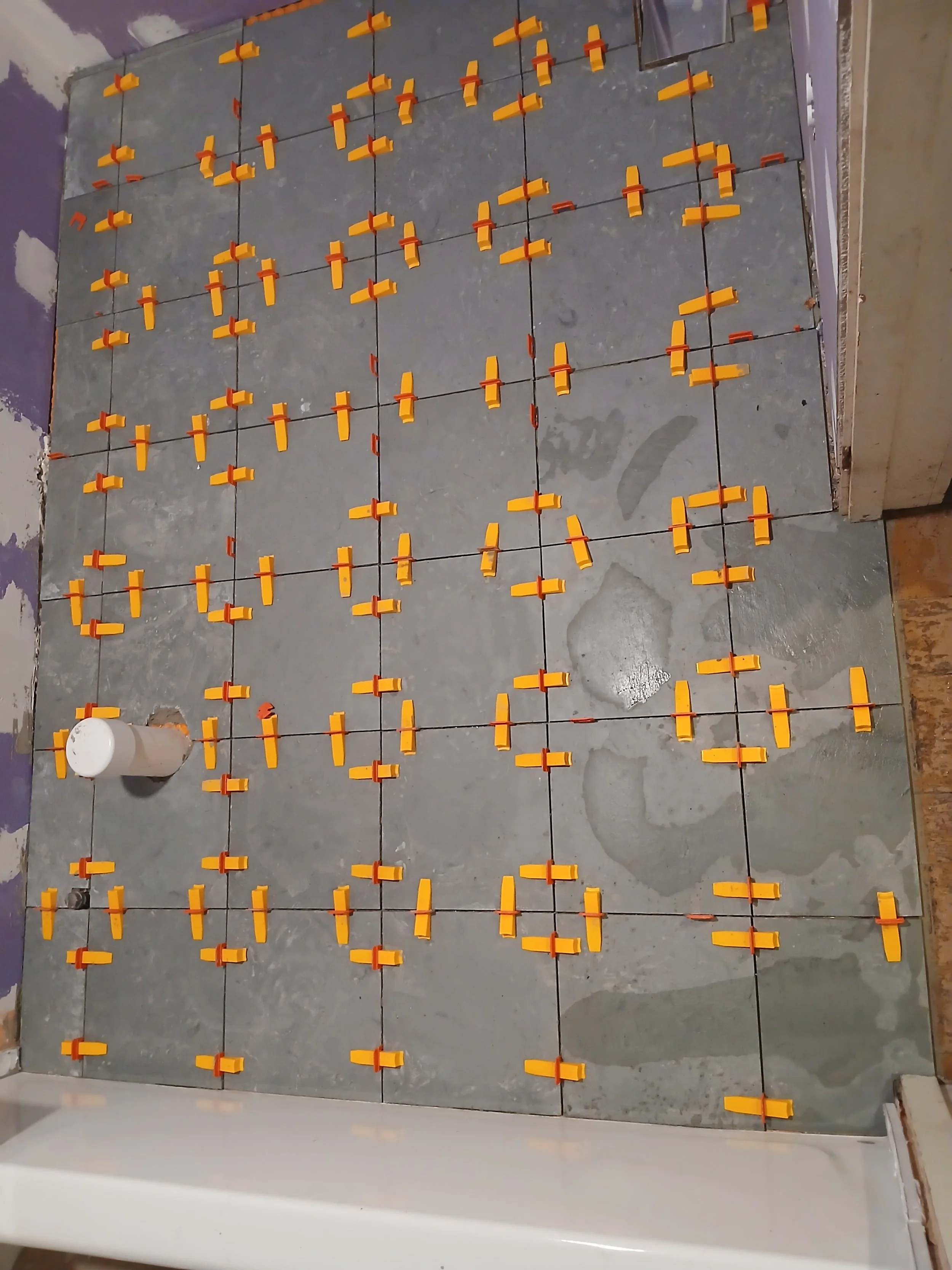 Floor being tiled with gray tiles, orange tile spacers evenly placed for grout lines, and a white bucket in the corner for tools or adhesive.