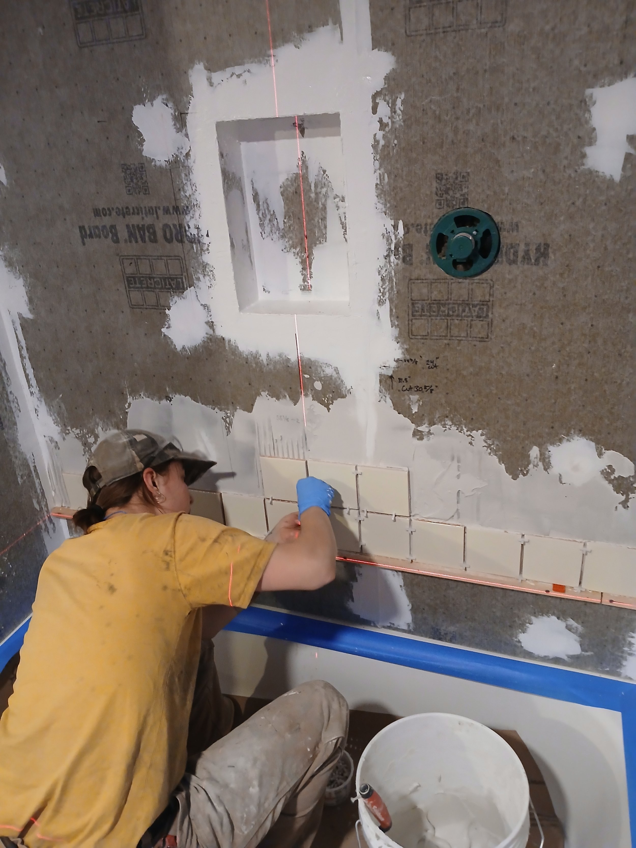 A person installing tiles on a wall with a laser level and a bucket of tile adhesive.