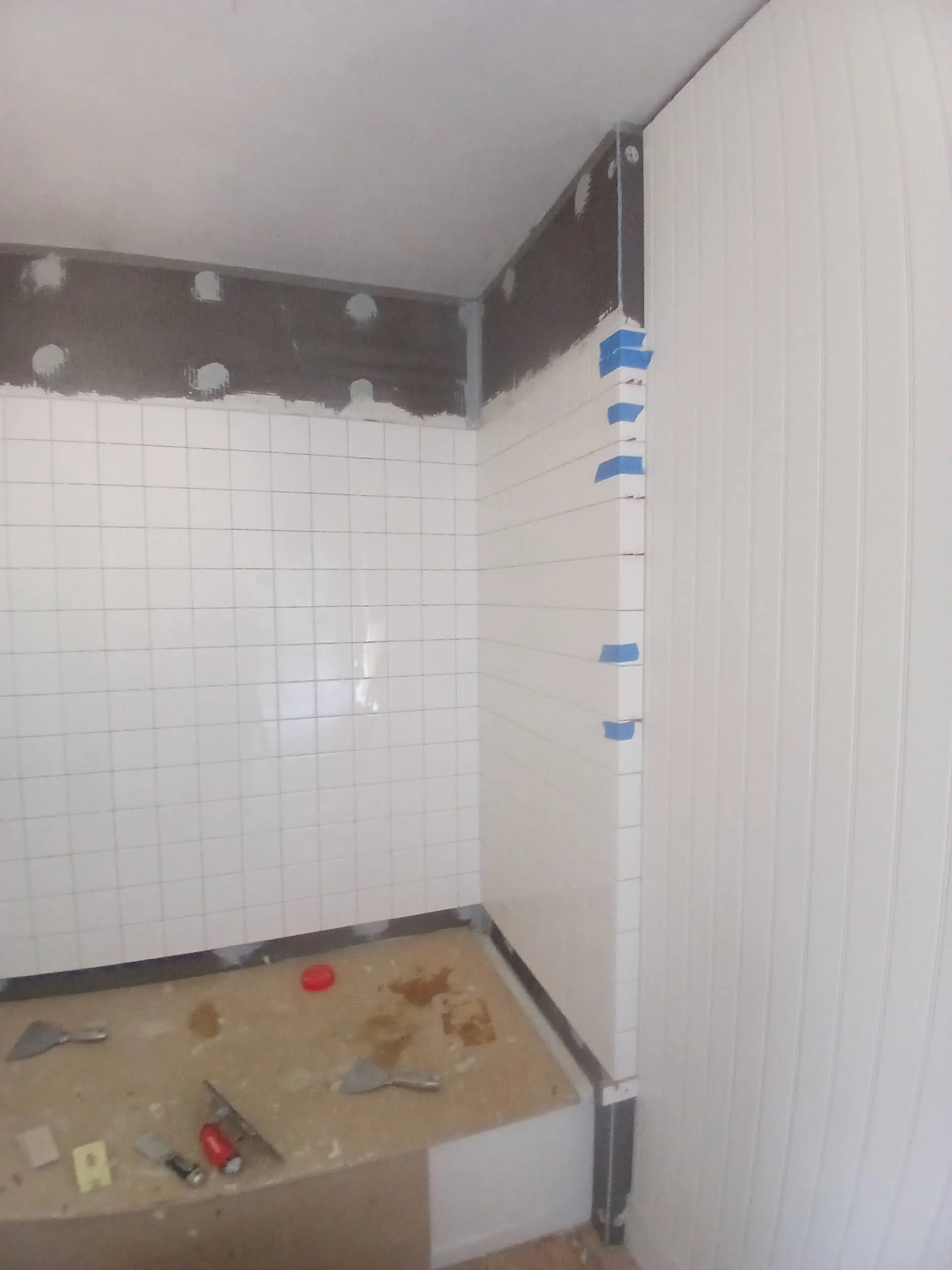 Bathroom under renovation with white tiles partially installed, wall tiles taped in place, tools on a makeshift work surface, and some construction materials.