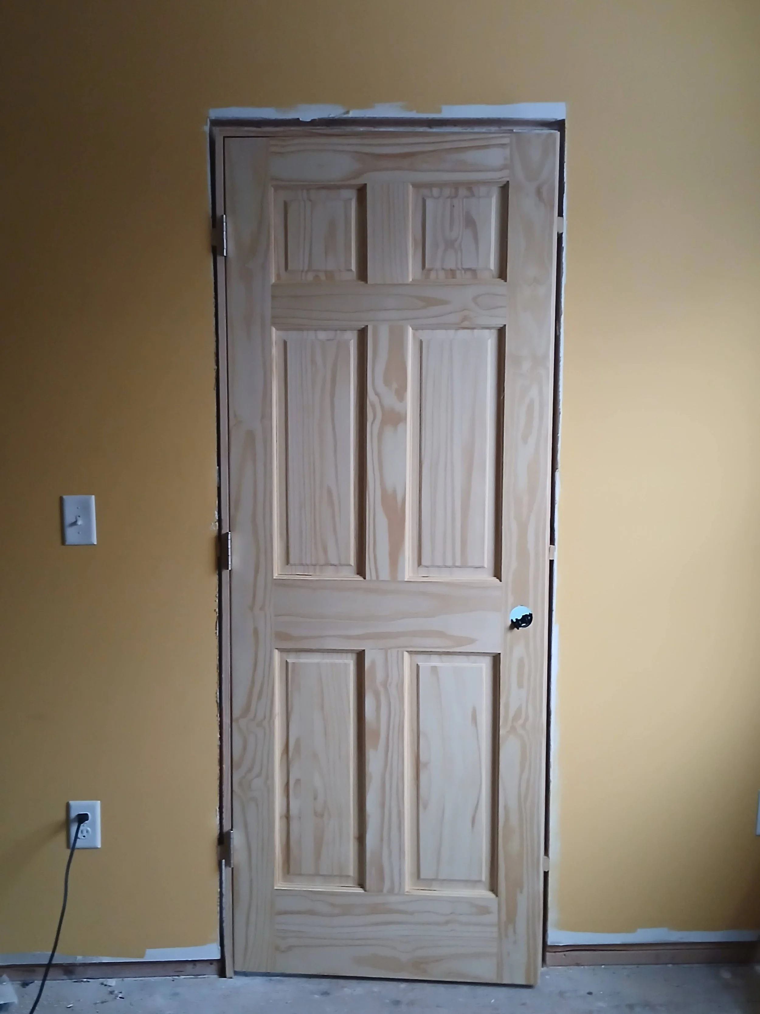unfinished six-panel wooden door leaning against a yellow wall in a room under construction or renovation.