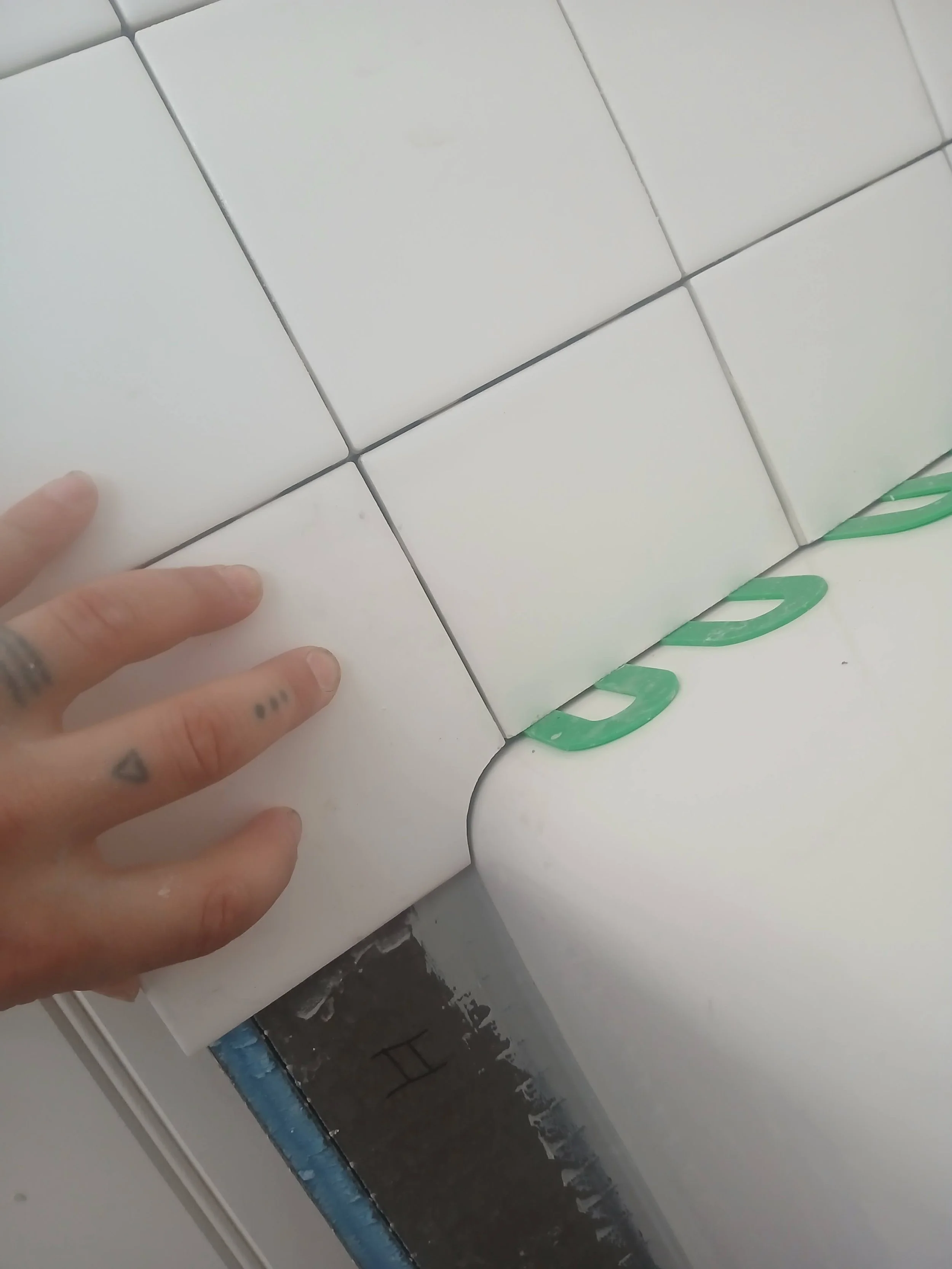 Close-up of a person's hand with tattoos touching white ceramic tiles and green tile spacers on a wall under construction.
