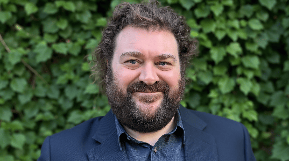 A professional male consultant with dark hair and beard, wearing a dark blue suit and shirt smiling at the camera