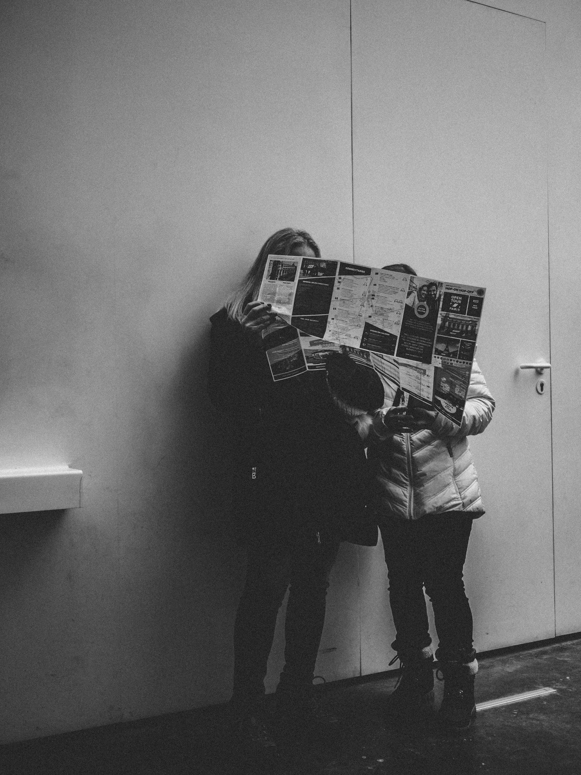 Two people standing against a plain wall, reading a large, colorful newspaper or brochure, with the person on the left partially obscured by it.