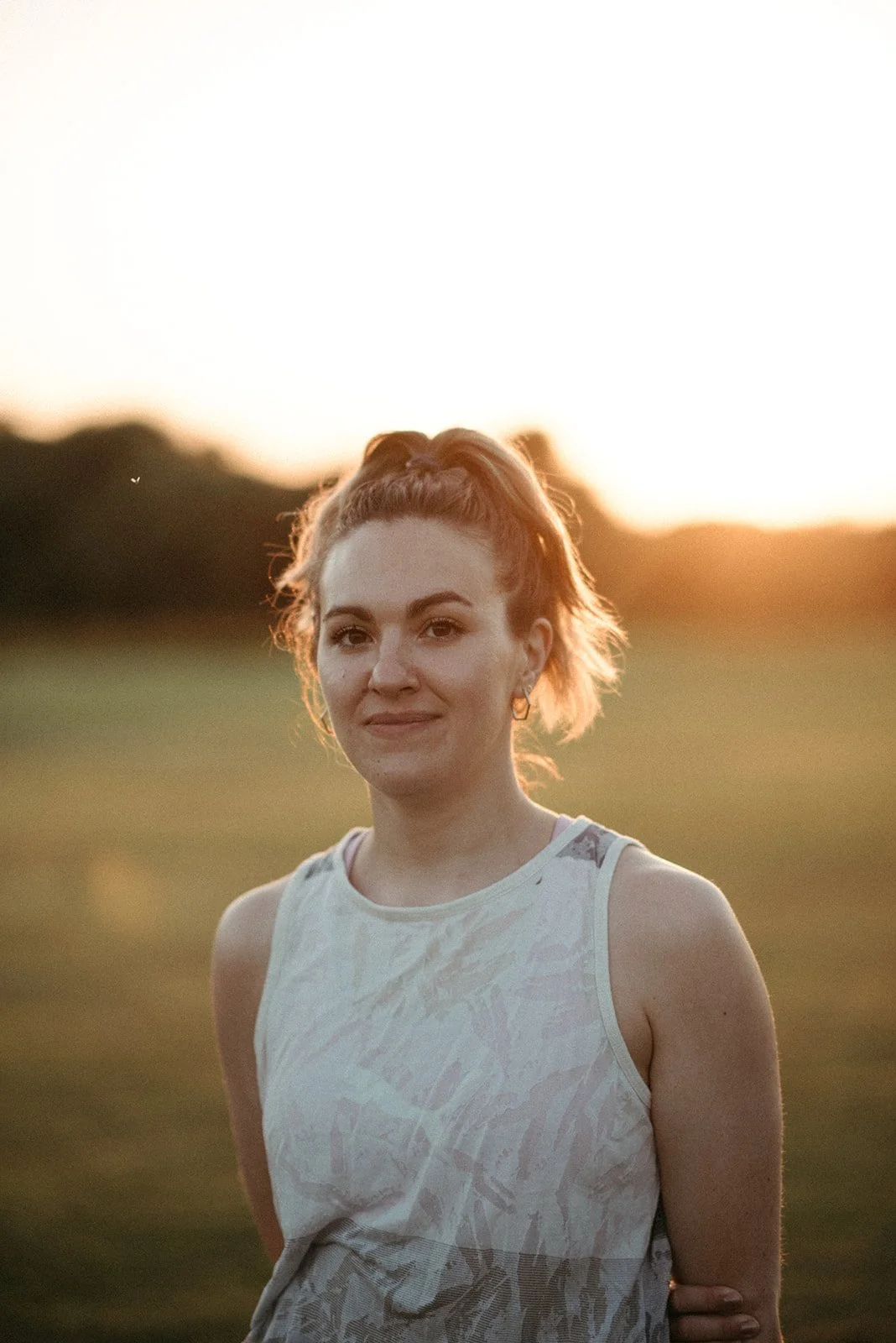A woman with light skin and reddish-brown hair tied in a high ponytail stands outdoors during sunset, wearing a sleeveless top with a subtle pattern, in a field with a blurred background of trees.