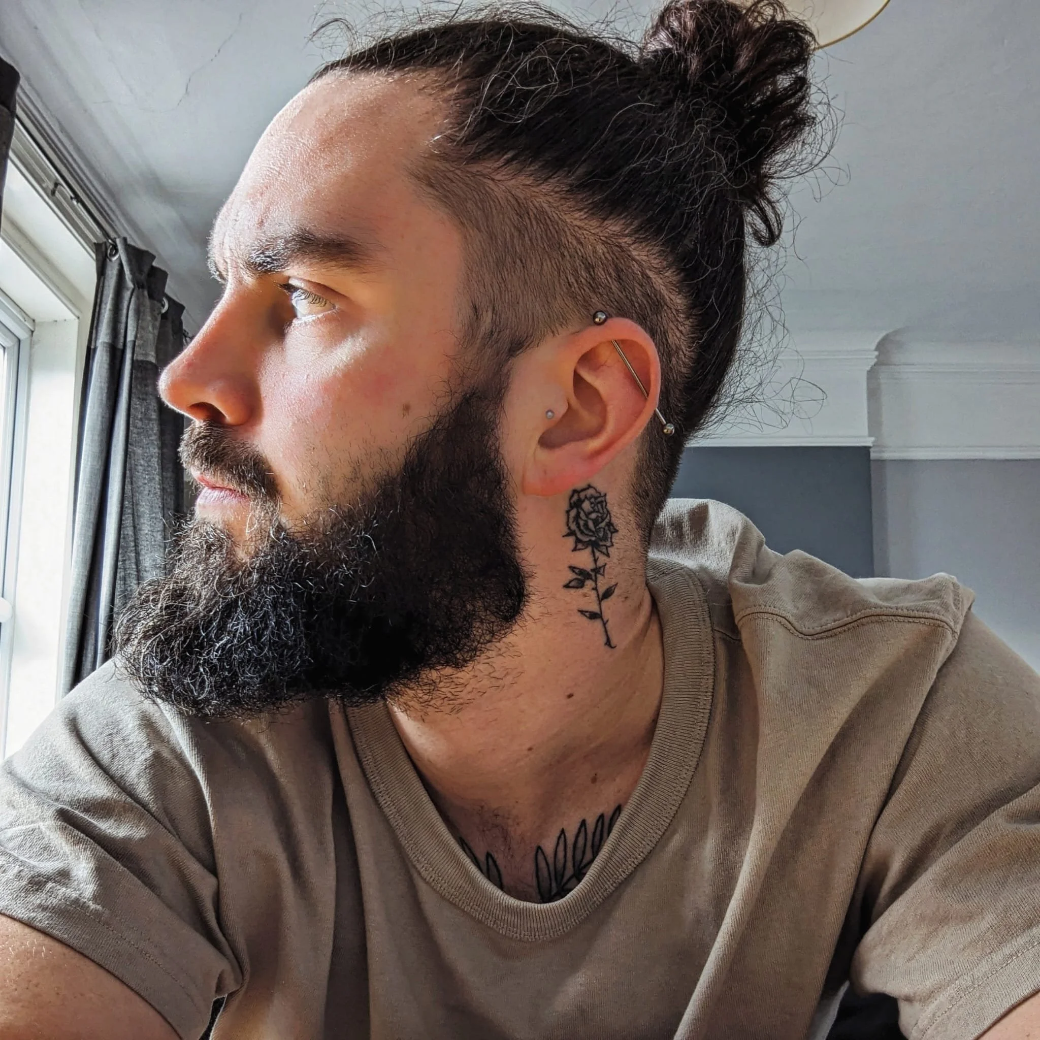A man with a beard looking out of a window, with ear piercings and a tattoo of a rose on his neck