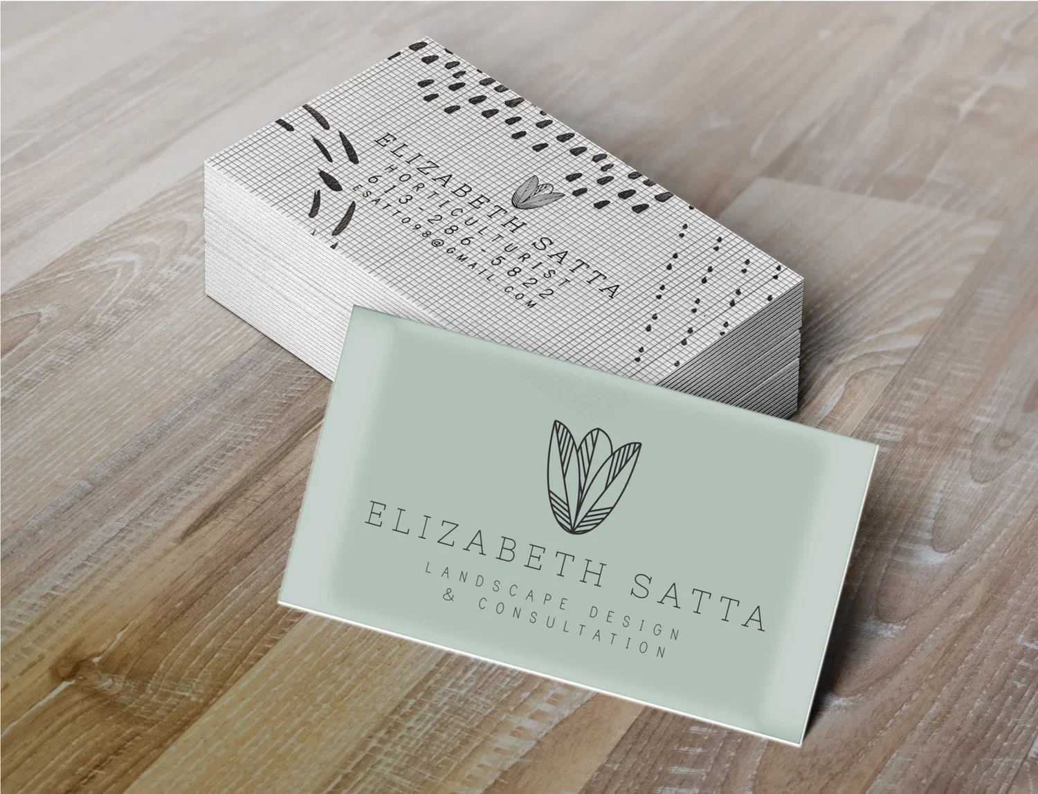 Business cards for Elizabeth Satta, a landscape design and consultation service, with a minimalist leaf logo on a wooden surface.