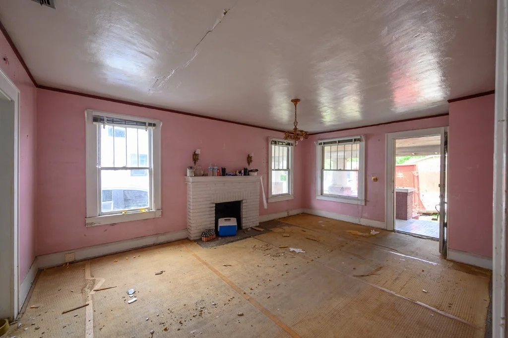 Living room with pink walls, a white brick fireplace, three windows, and a ceiling with a crack. The room is empty with some debris on the floor and an open door leading outside.