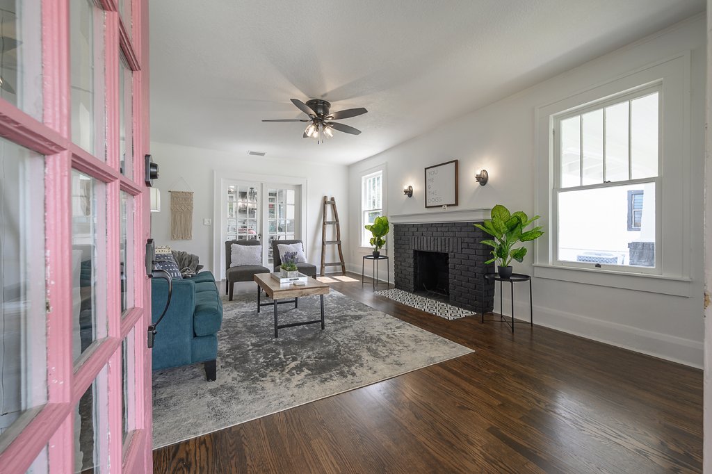 Living room with a pink-framed glass door, a dark teal sofa, and gray armchairs surrounding a wooden coffee table. There is a brick fireplace with two potted plants on black stands, a framed quote, and large windows letting in natural light. The room has wooden floors and a gray and white rug.