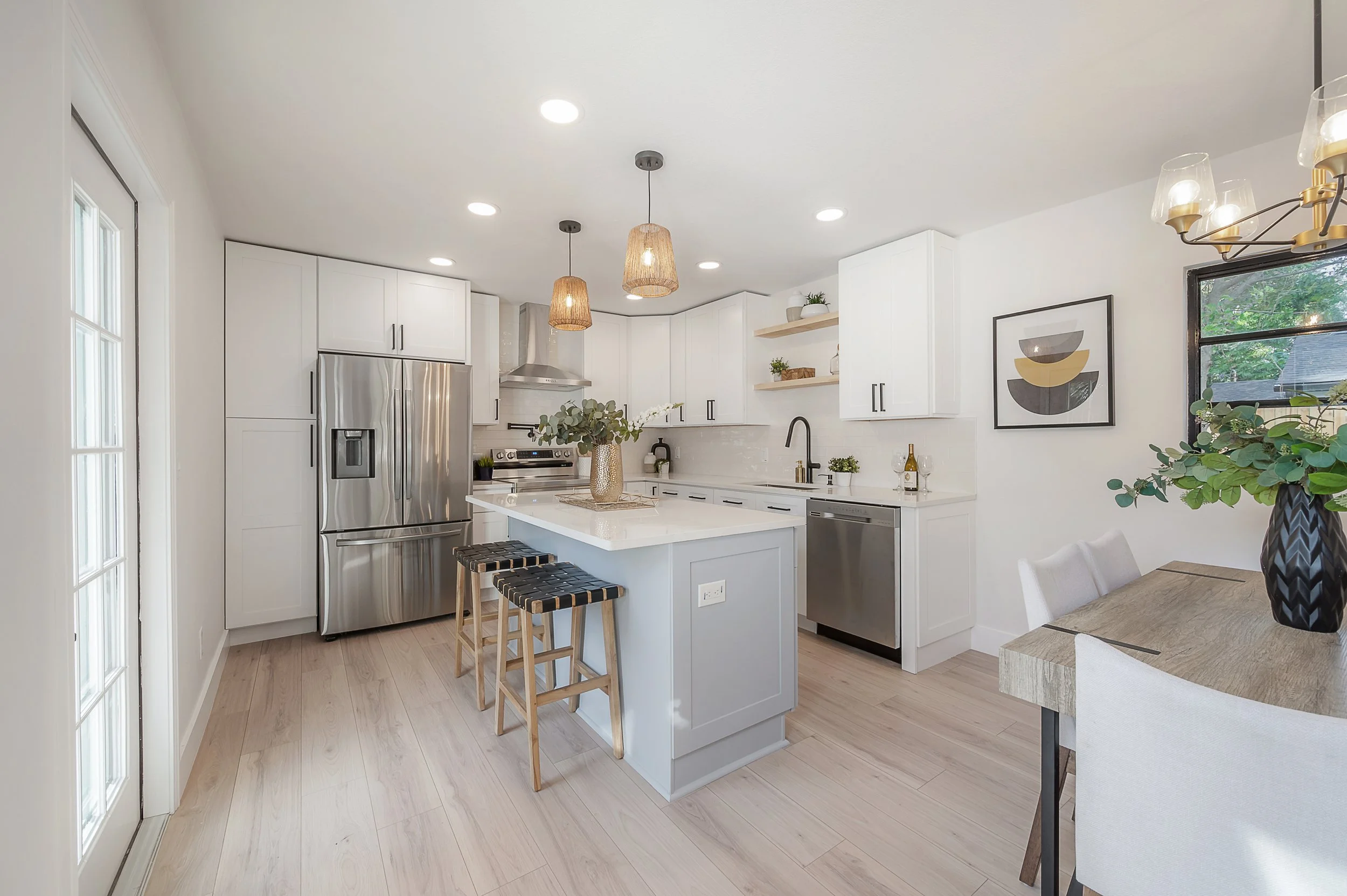 Bright modern kitchen with white cabinets, a central island with seating, stainless steel appliances, open shelves with decor, and a dining area with a wooden table and plants.
