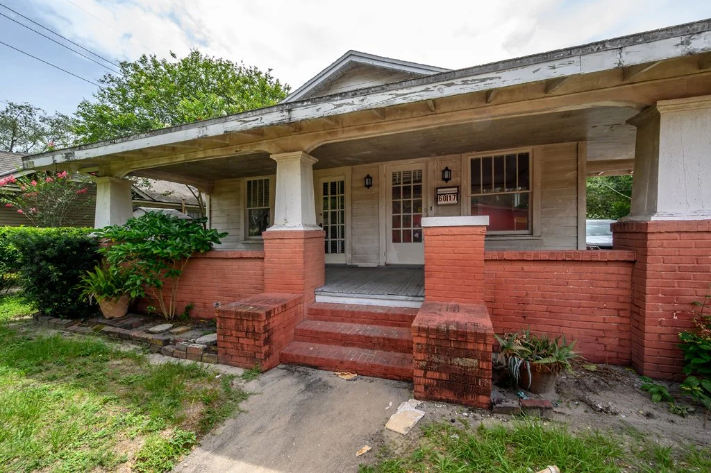 Front porch of a house with brick steps, white columns, and a wooden porch ceiling. There are plants on each side of the steps, and the house has white siding with large windows. The house appears to be slightly aged.