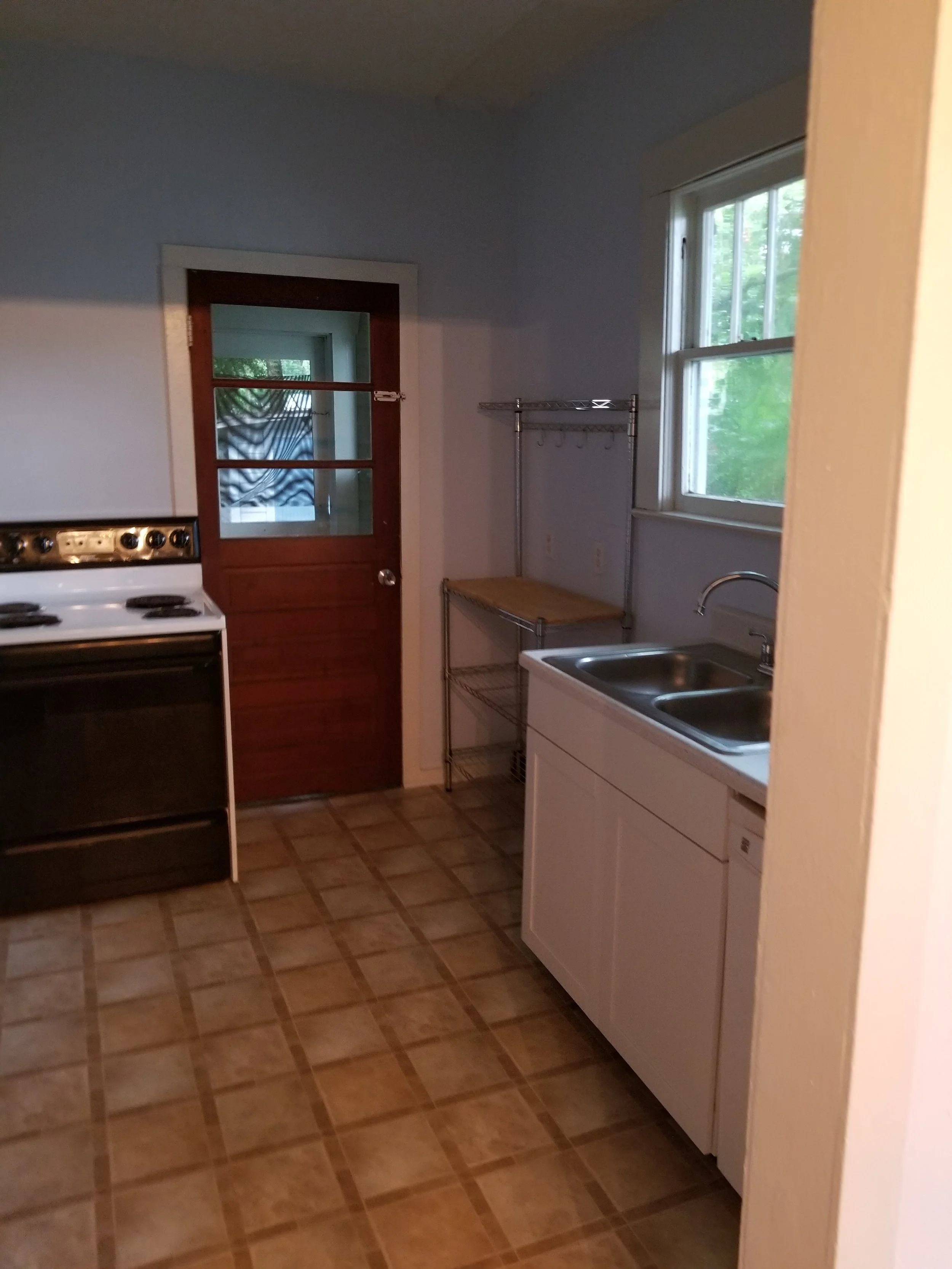 Small kitchen with a white sink, a window, a wooden door with window panes, a black stove, and a metal shelving unit, with tiled flooring.
