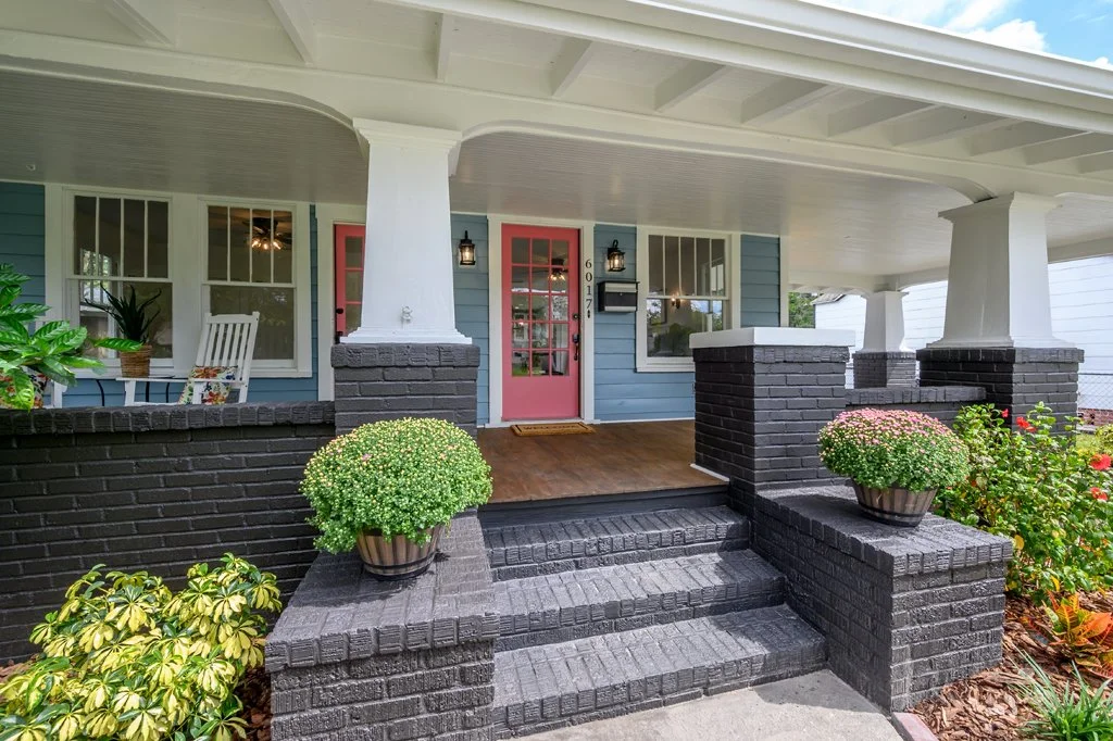 Front porch of a house with black brick stairs, two potted plants, and a pink front door with glass panels, located in a neighborhood with other houses visible in the background.