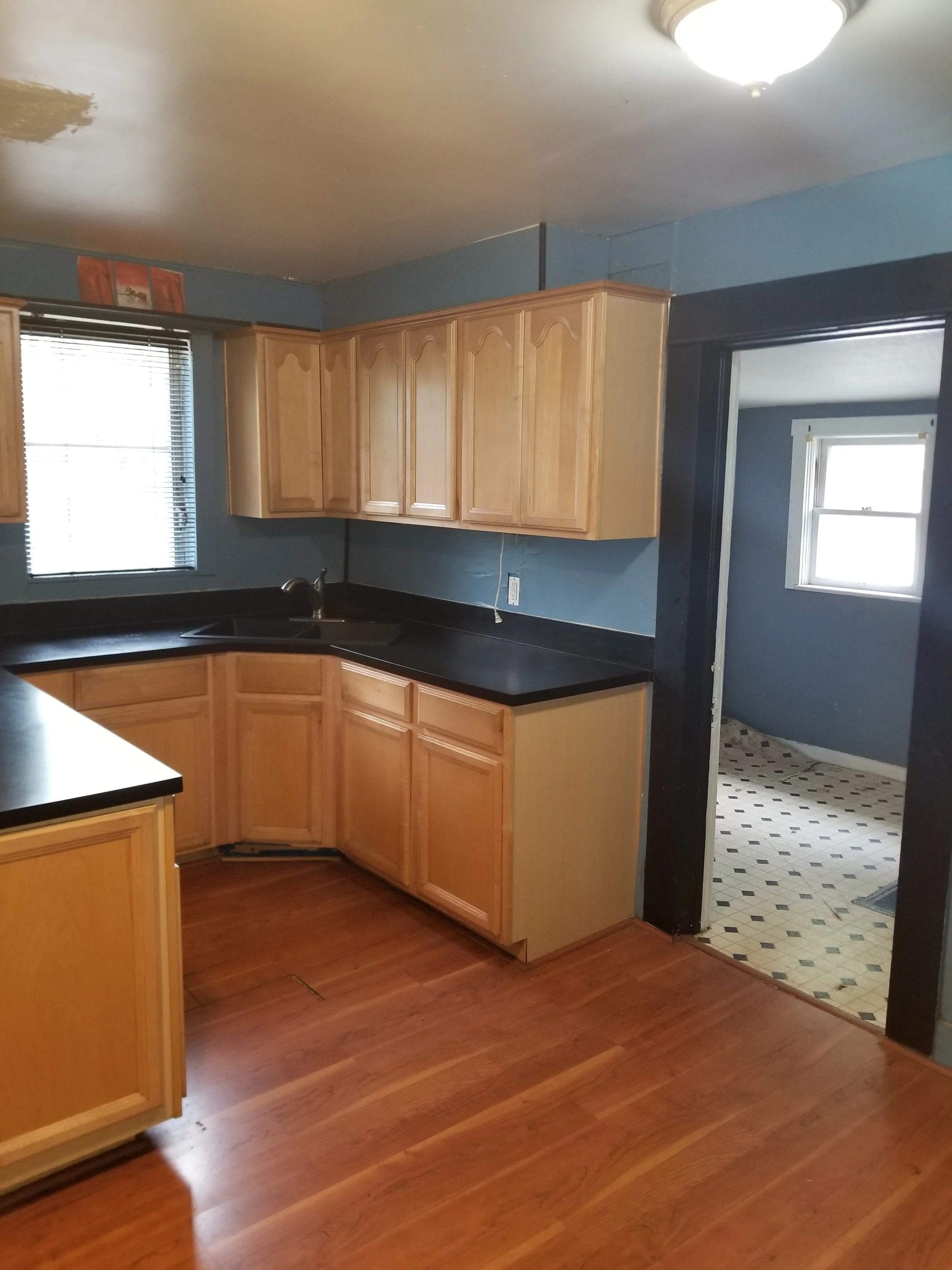 Kitchen with wooden cabinets, black countertops, a window with blinds, and a doorway leading to a room with patterned linoleum flooring and a small window.