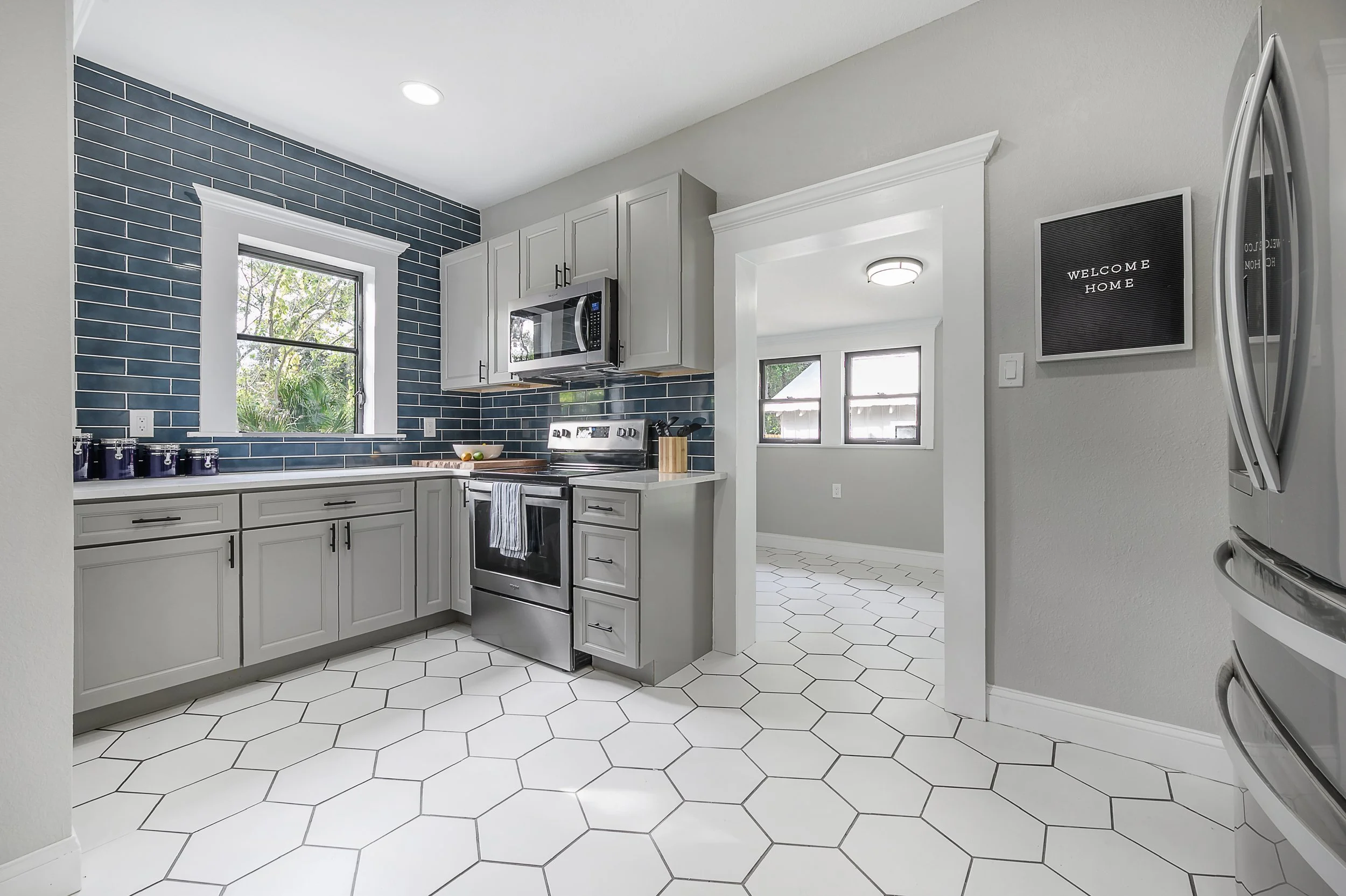 A modern kitchen with white cabinets, stainless steel stove and microwave, dark blue subway tile backsplash, hexagonal white floor tiles, gray walls, and a window looking outside.