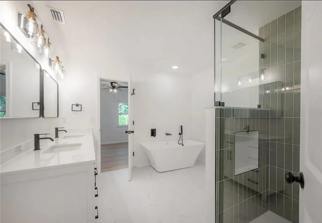 Modern bathroom with white vanity, black faucet, double mirrors, walk-in shower with green tiles, and freestanding bathtub, with doorway leading to a room with a ceiling fan.