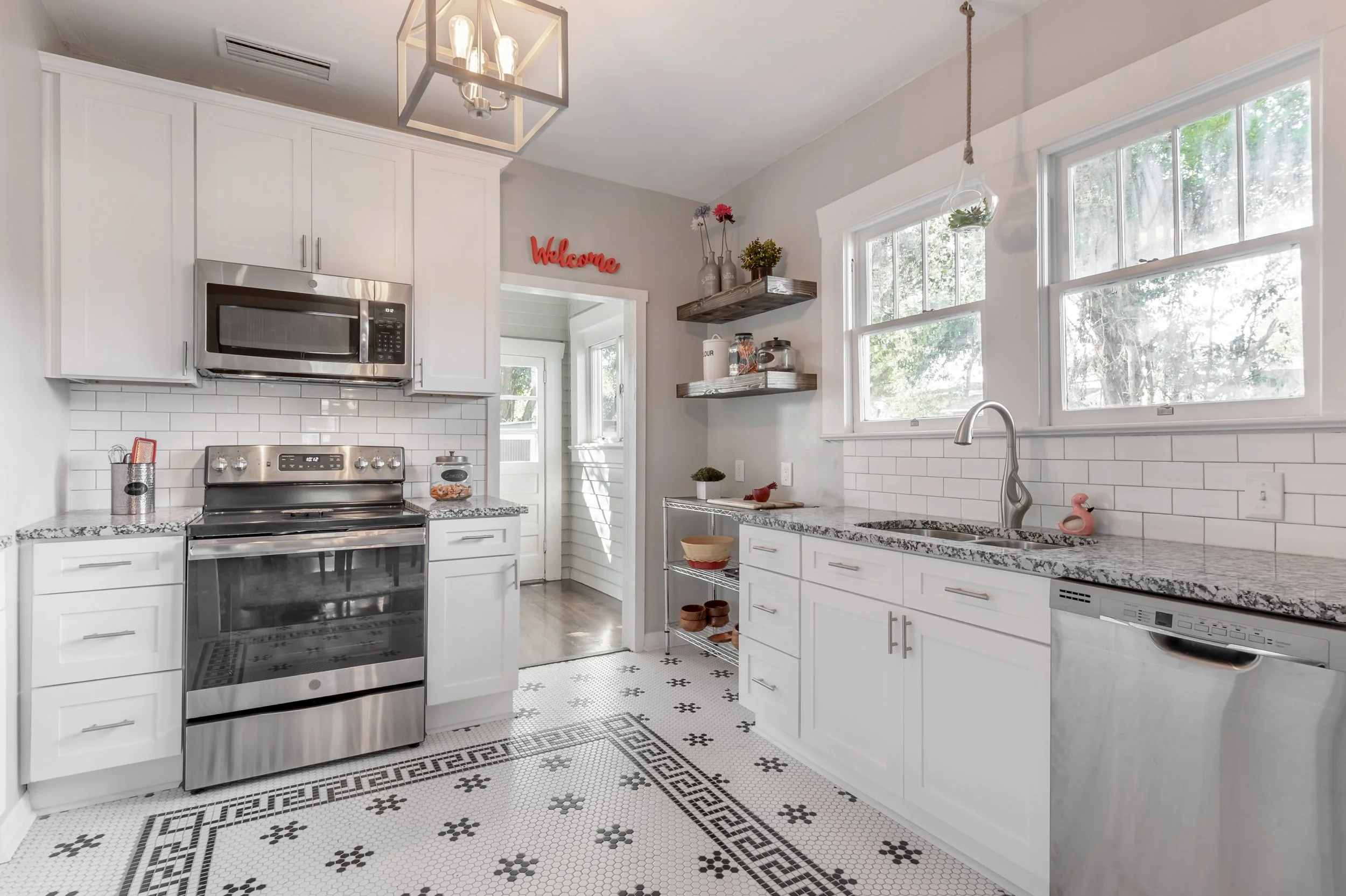 A modern kitchen with white cabinetry, a gray granite countertop, stainless steel appliances, and black and white patterned tile flooring. There are windows above the sink, open shelving with decorative containers, and a small pink flamingo figurine on the countertop.