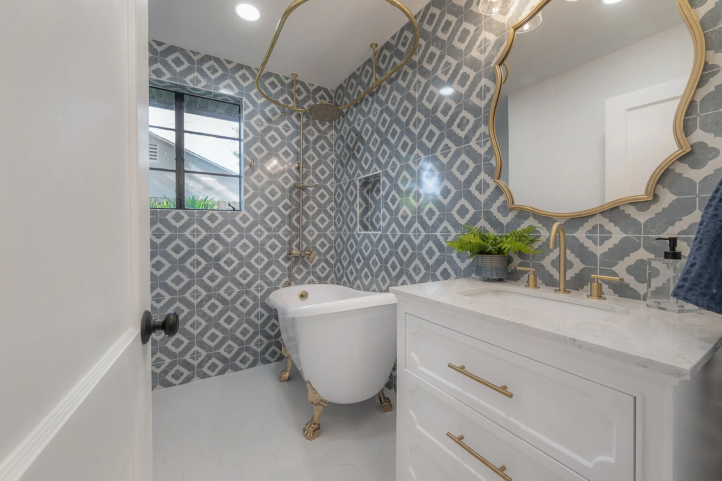 Bathroom with patterned gray and white tile walls, a freestanding bathtub with clawfoot legs, a vanity with a marble countertop and gold fixtures, a decorative mirror, a potted plant, and a small window.