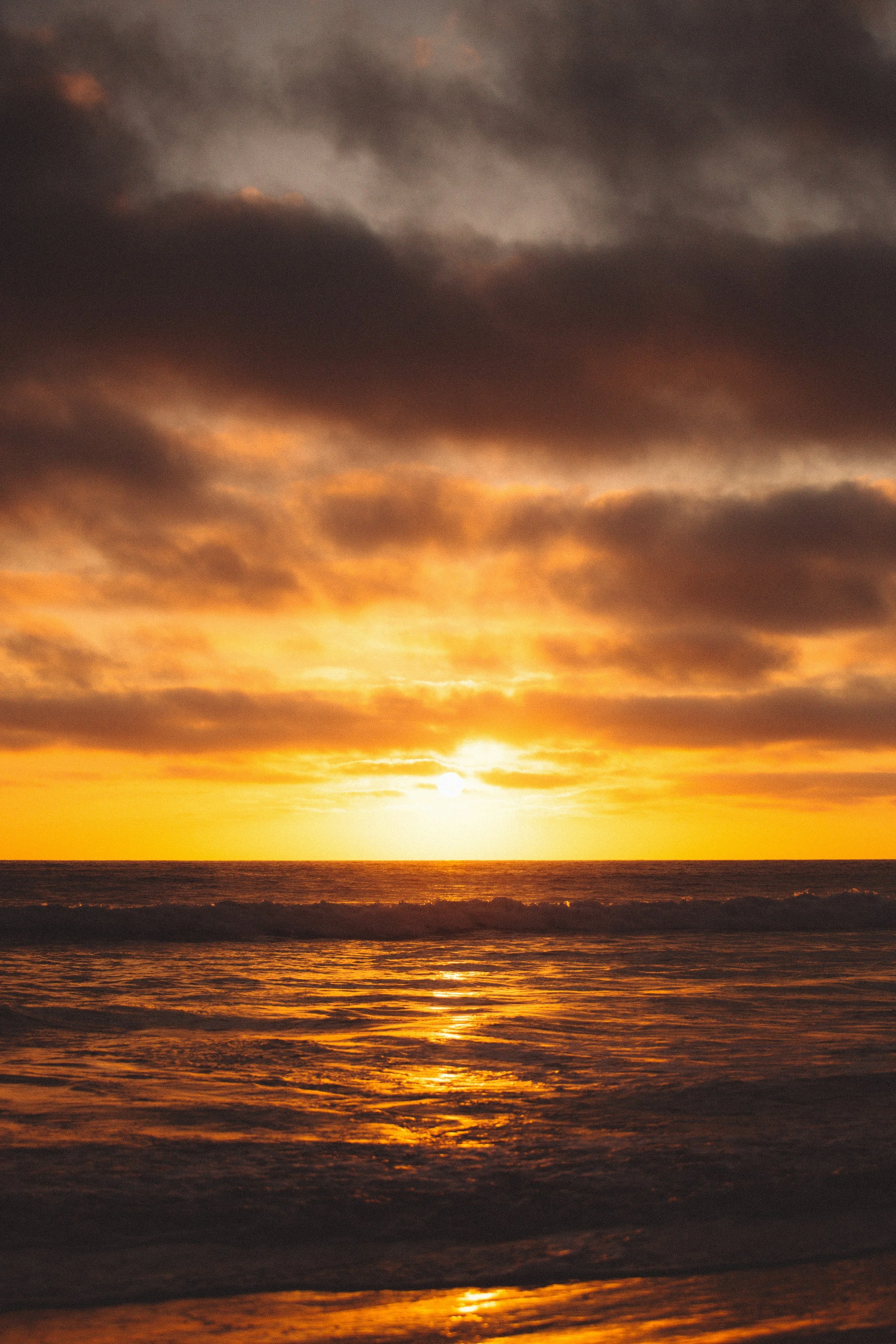 Sunset over the ocean with dark clouds and reflected orange and yellow hues on the water.
