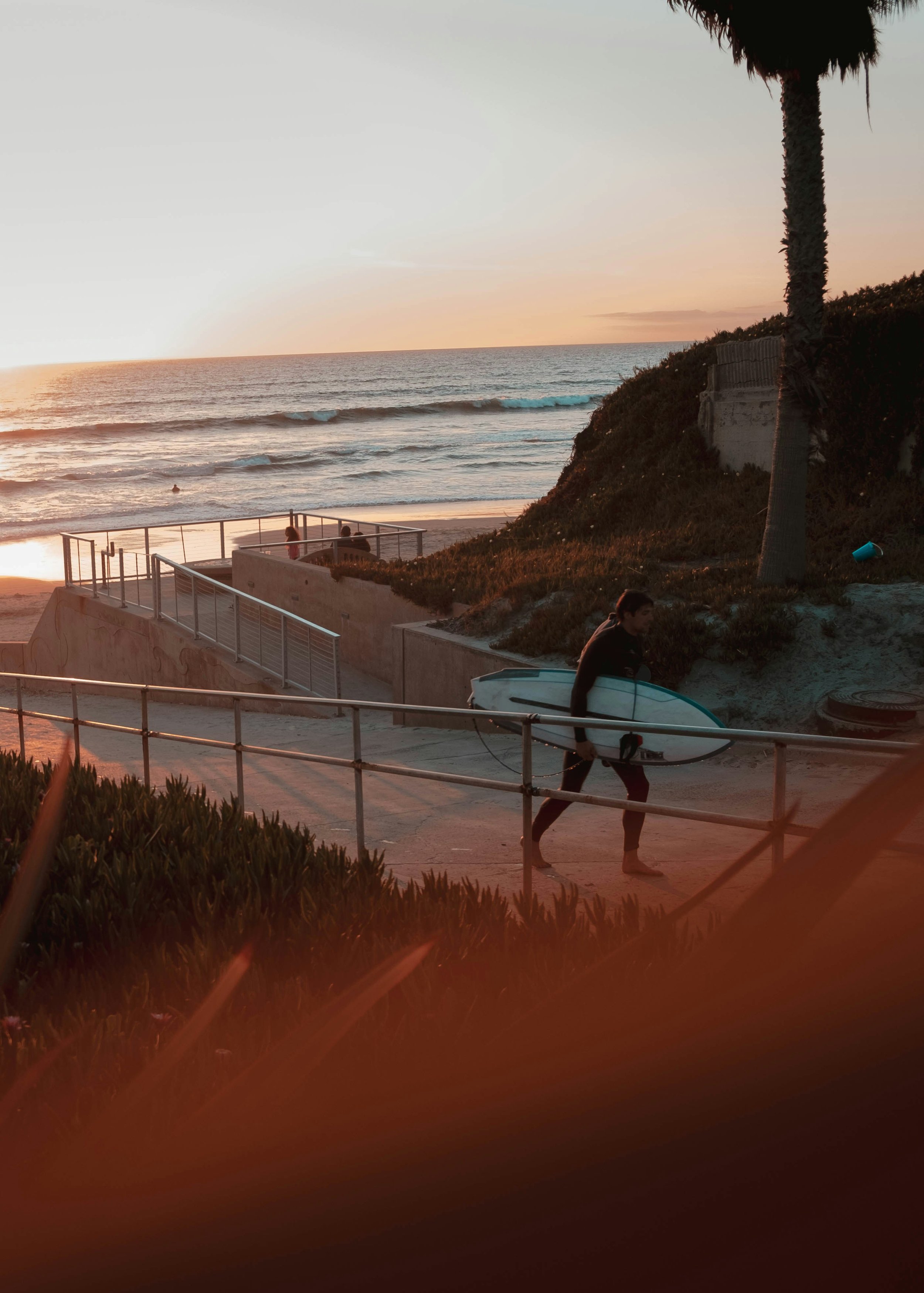 A person walking with a surfboard along a pathway near the beach at sunset, with a palm tree, ocean waves, and a few people in the distance.