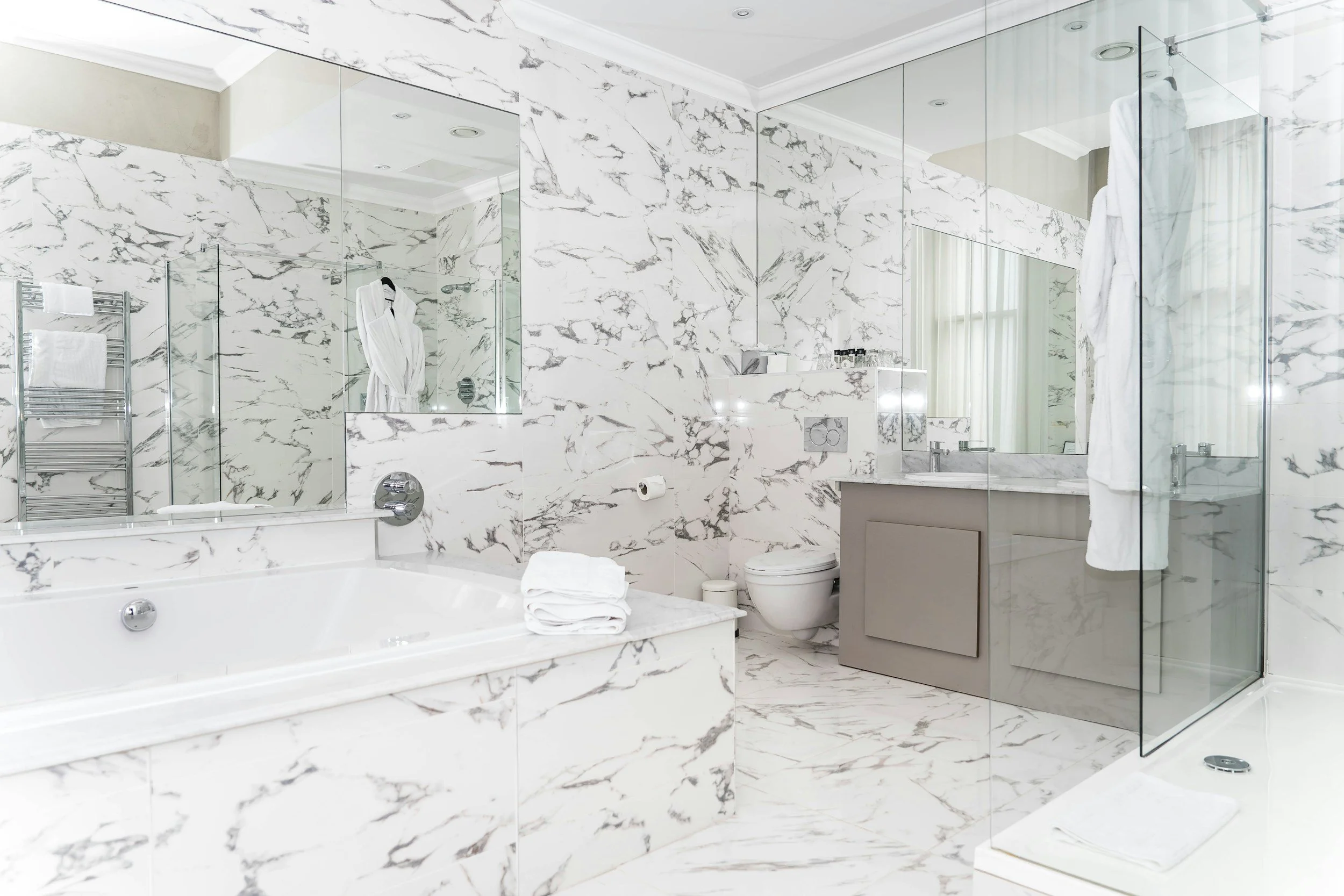 Modern bathroom with extensive white marble walls and flooring, a bathtub with towels, a toilet, a glass-enclosed shower, and a vanity with a large mirror.