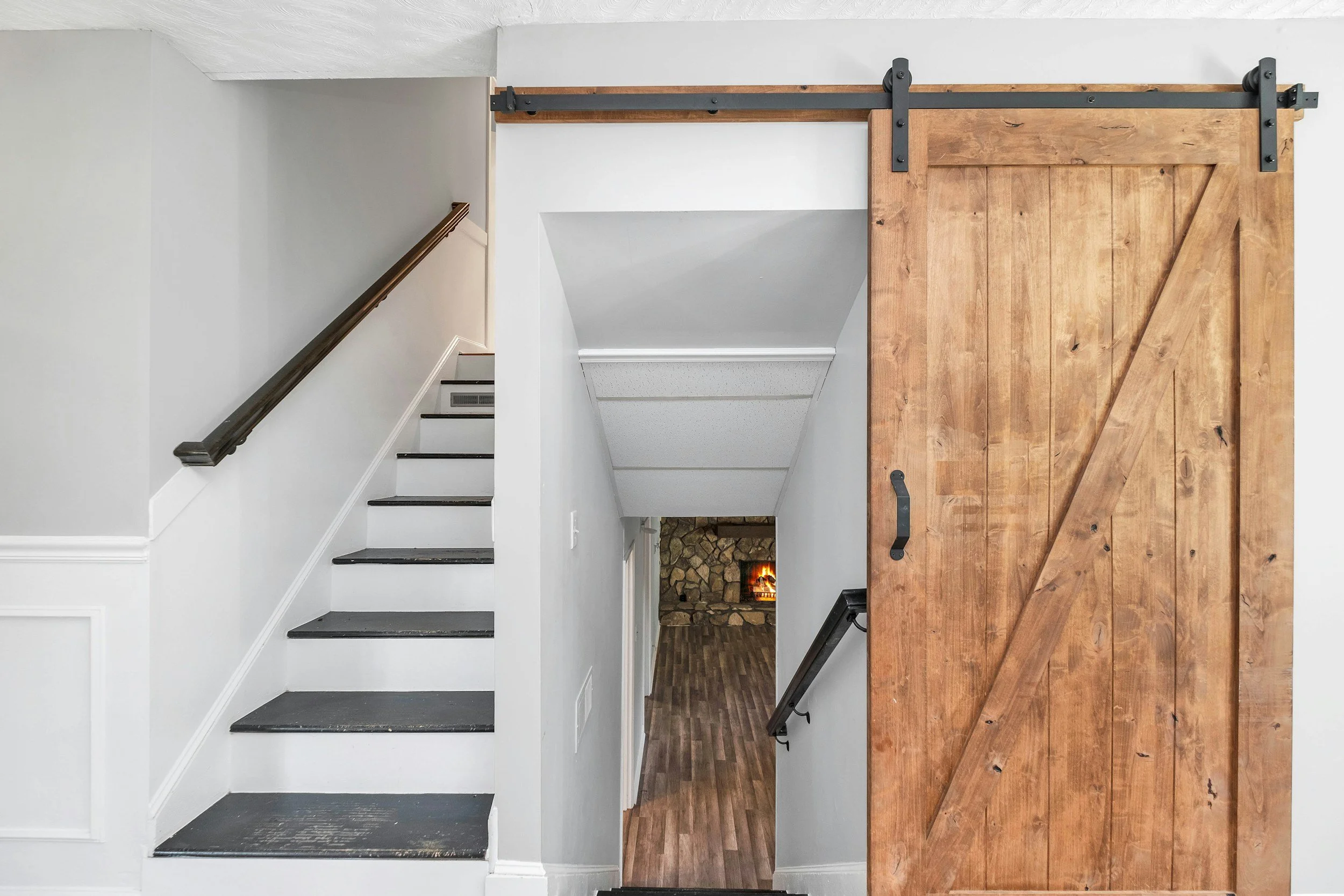 Interior view showing a staircase with white walls, black steps, and a wooden sliding barn door with black hardware, leading to a room with a fireplace.