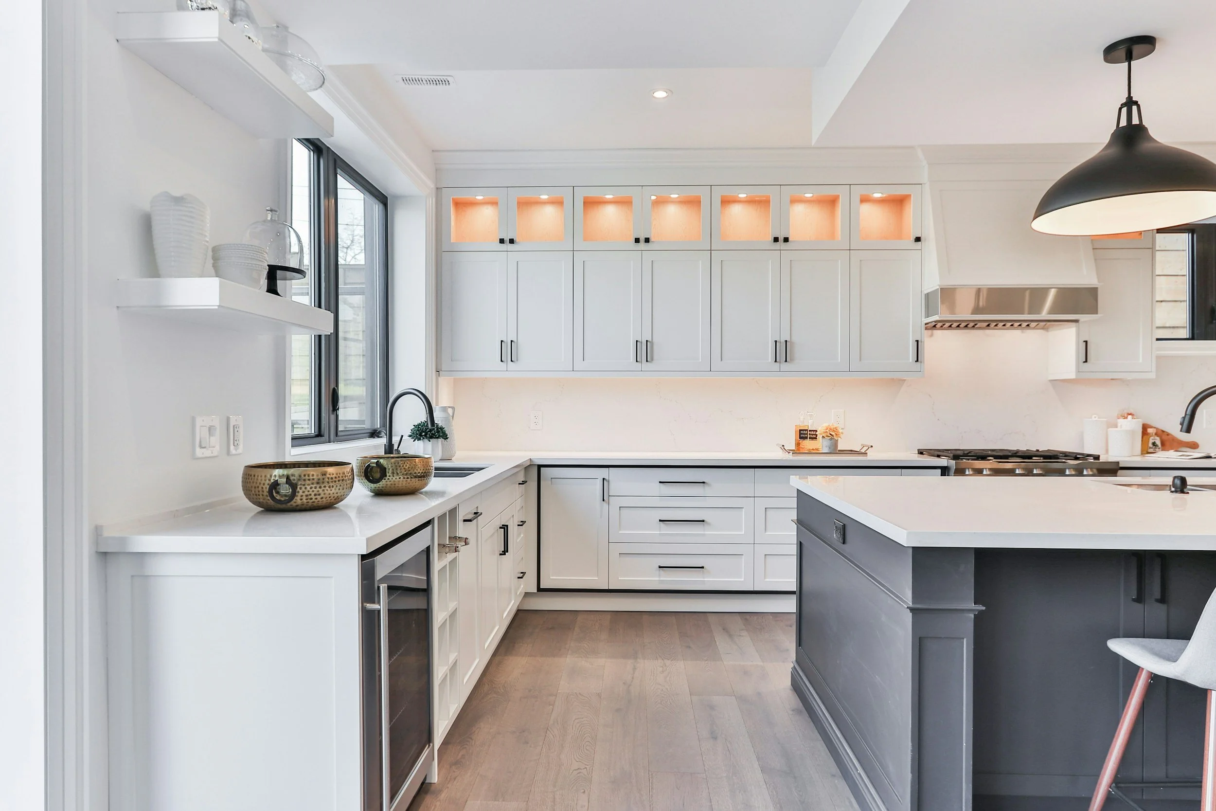 Modern kitchen with white cabinets, a gray island, and wood flooring. Features include a window, black handles on cabinets, a black pendant light, and decorative bowls on the counter.