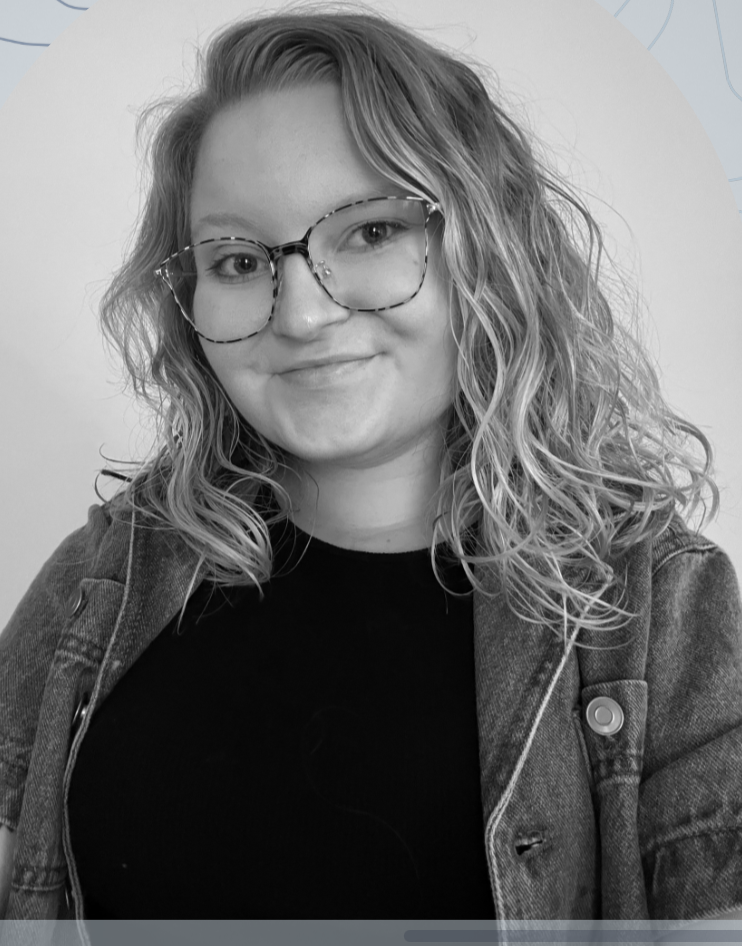 A young woman with wavy blonde hair, wearing glasses and a jean jacket, smiling outdoors.