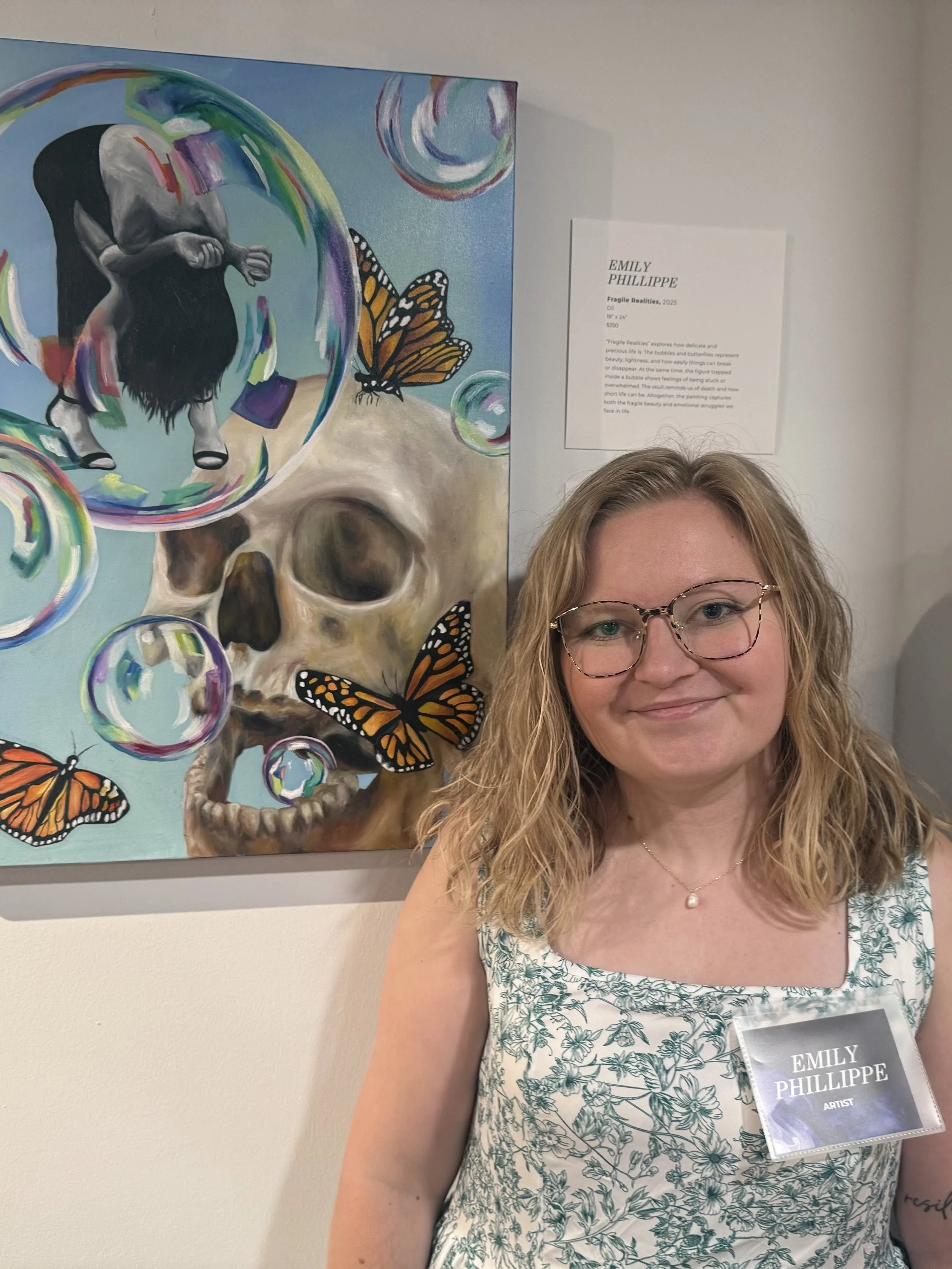 A woman with curly blonde hair, glasses, and a pearl necklace smiling in front of a colorful painting of a skull, children and butterfly, displayed at an art exhibit. She is wearing a sleeveless dress with a green floral pattern and has an artist badge reading 'Emily Philippe'.