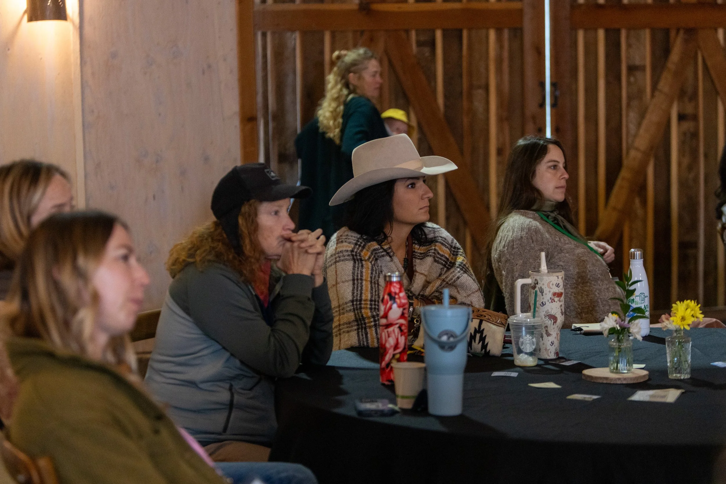 A group of women sitting at a table in a rustic barn-like setting, attentively listening, with drinks and flowers on the table.