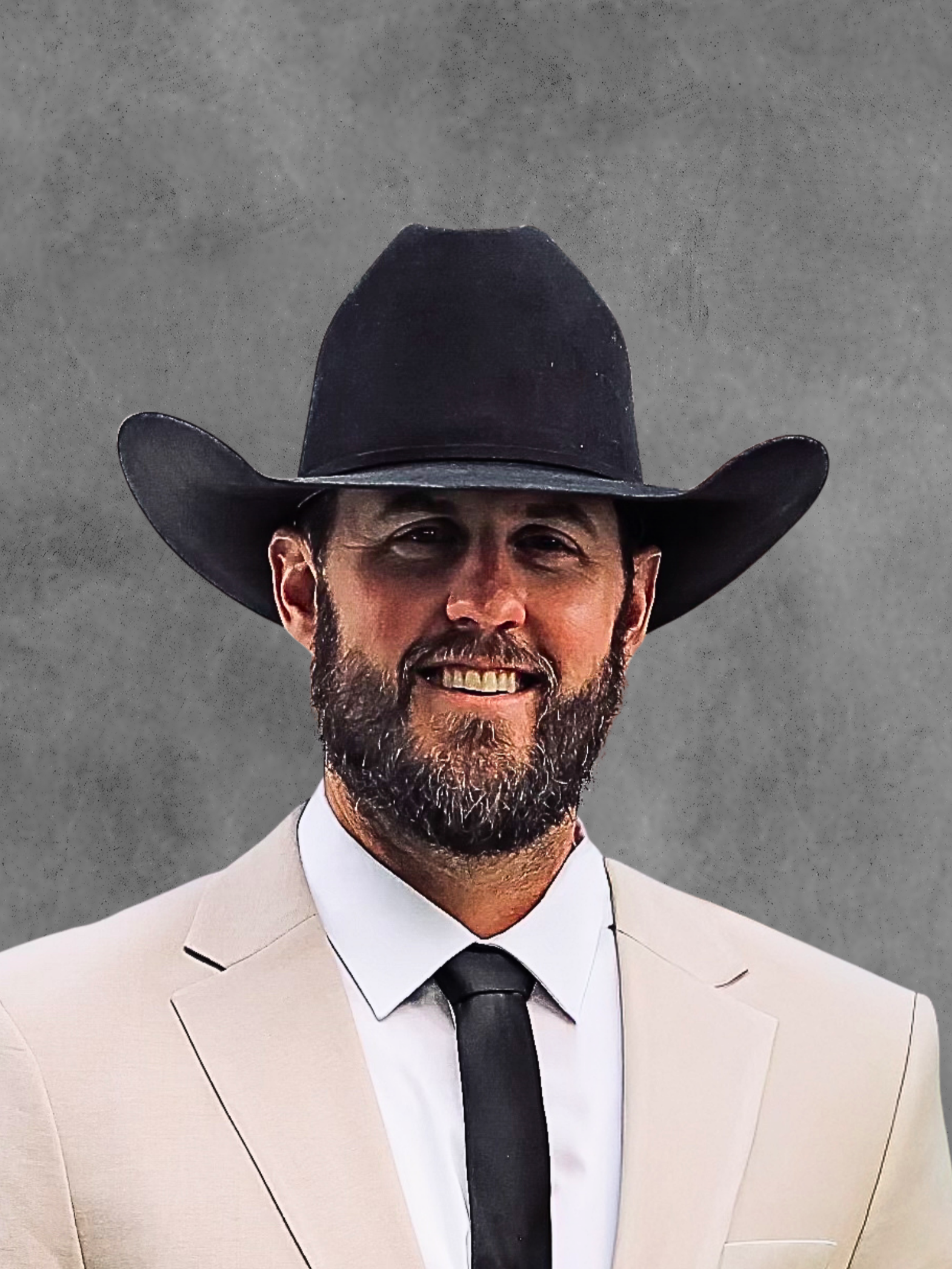 A man wearing a black cowboy hat, sunglasses, a beige suit jacket, white shirt, and black tie, smiling against a gray background.