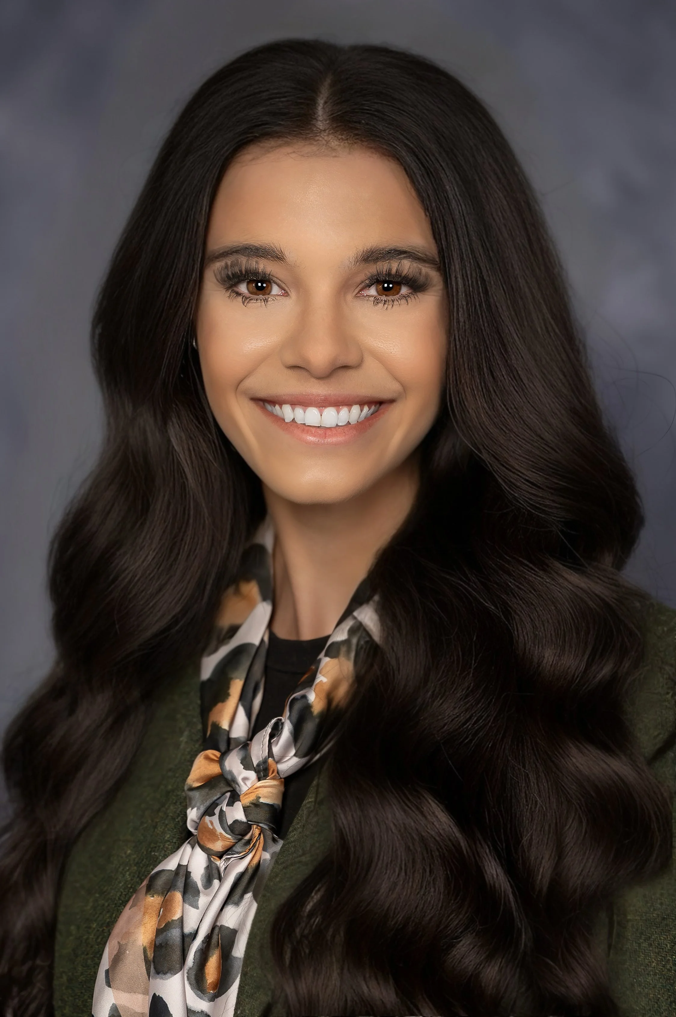 A woman with long dark hair smiling, wearing a patterned scarf and a green top, against a gray background.