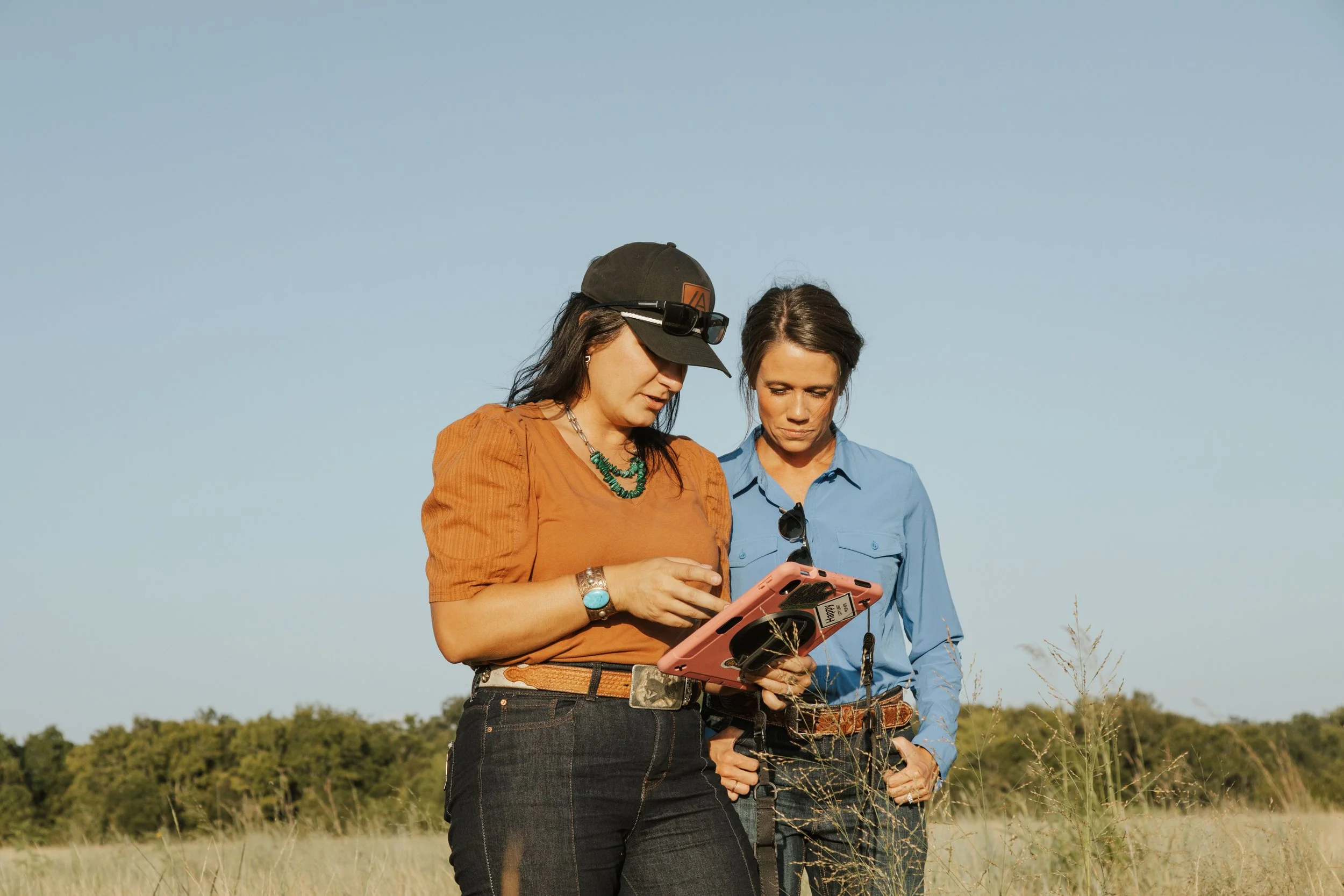 Two women standing outdoors in a grassy field, looking at a smartphone together, with a clear blue sky above and trees in the background.