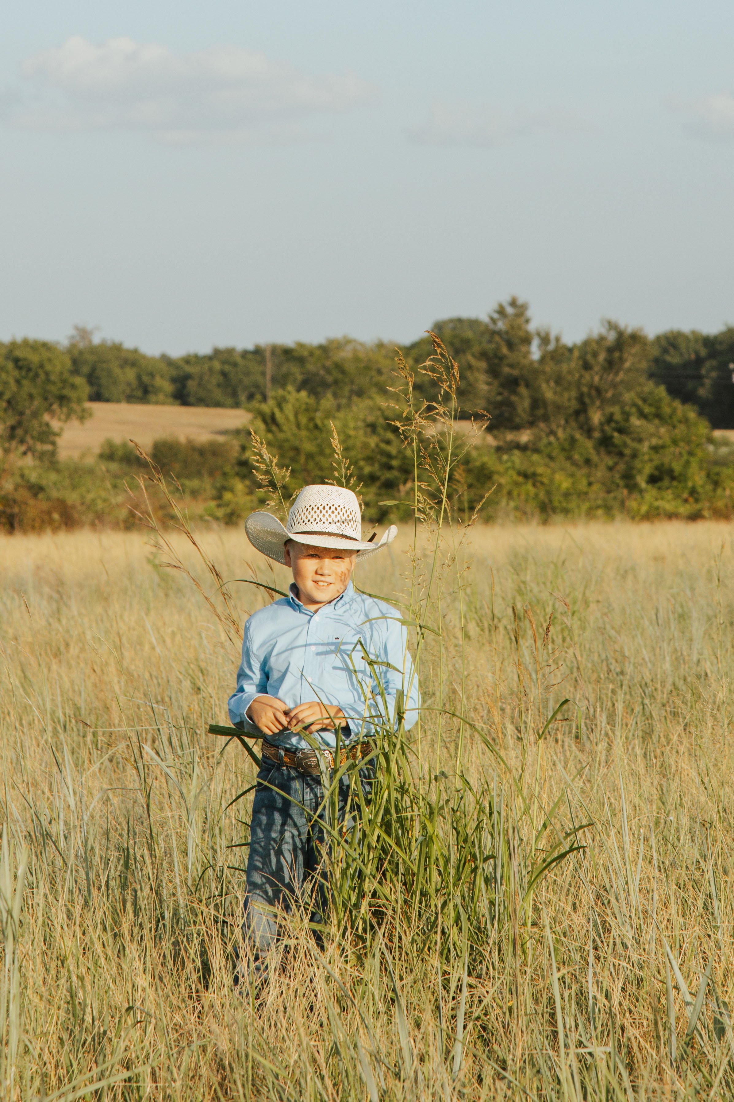Young boy in a cowboy hat standing in tall grass in a field, smiling, with trees and a partly cloudy sky in the background.