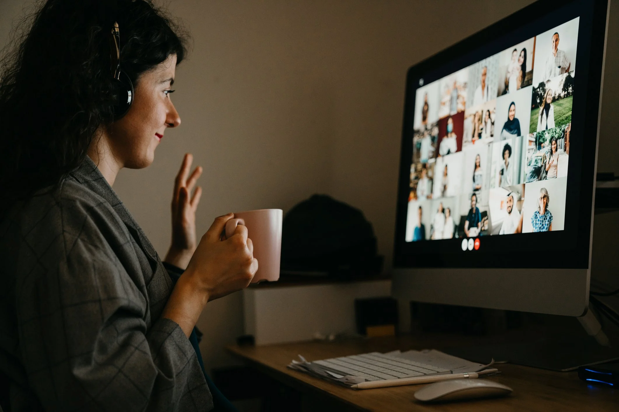 Woman sitting at a desk on a video call with multiple participants, holding a coffee mug, waving, wearing headphones, and looking at her computer screen with a collage of faces.