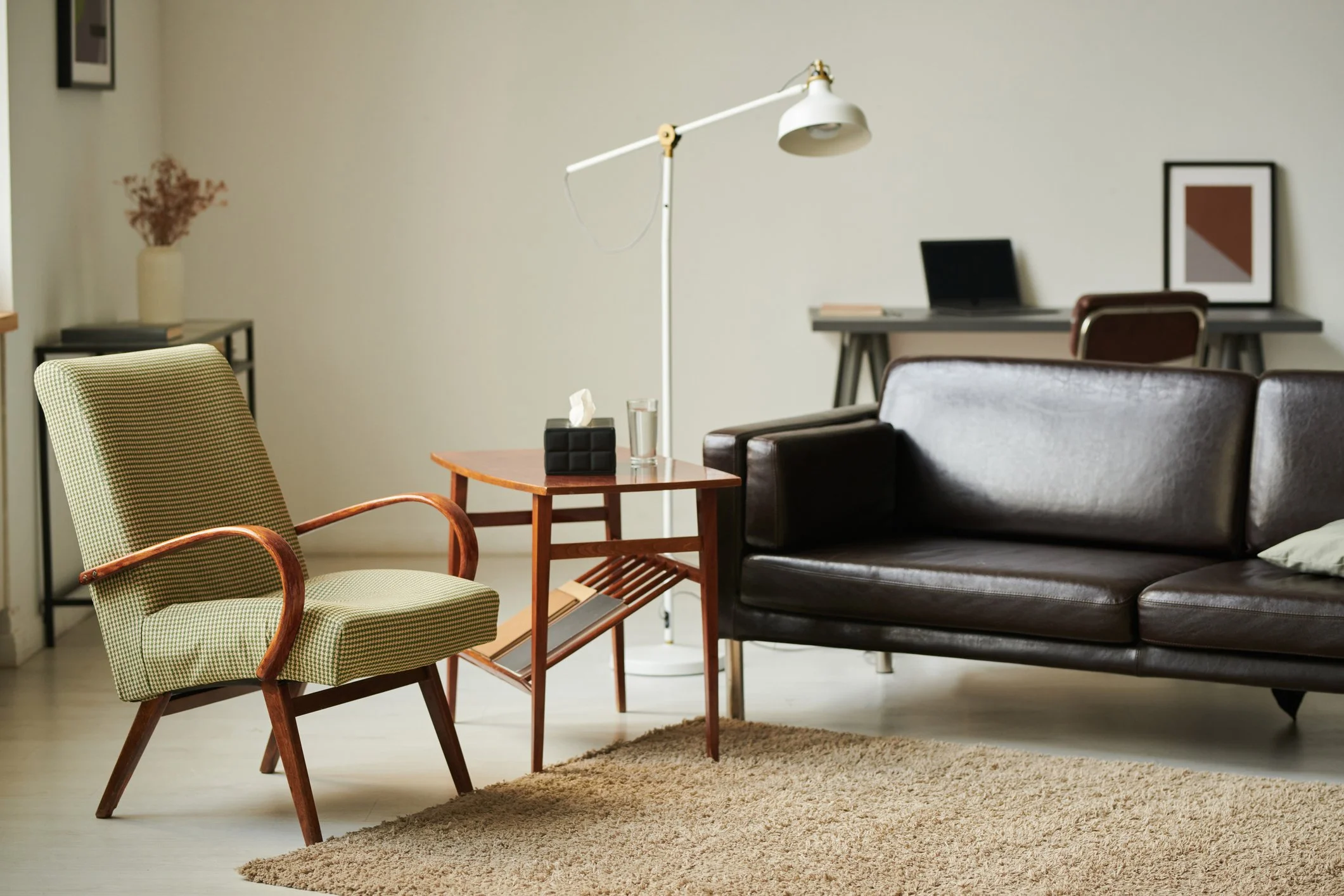 Living room with green armchair, dark brown leather sofa, white floor lamp, small wooden side table, beige rug, and desk with laptop and picture frame on wall.