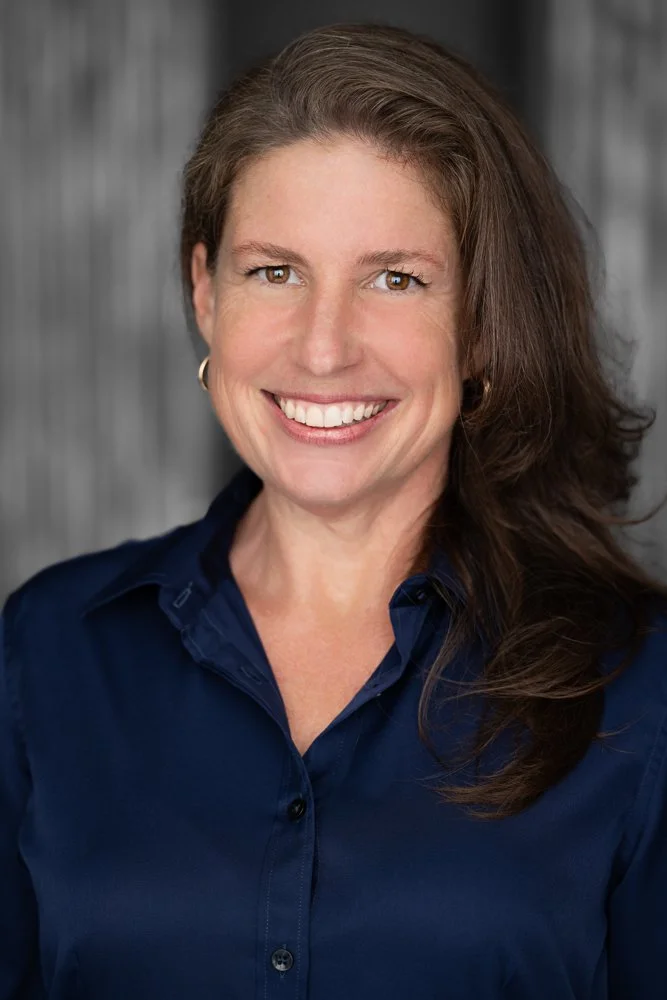 A smiling woman with long brown hair wearing a navy blue shirt standing in front of a gray wooden background.