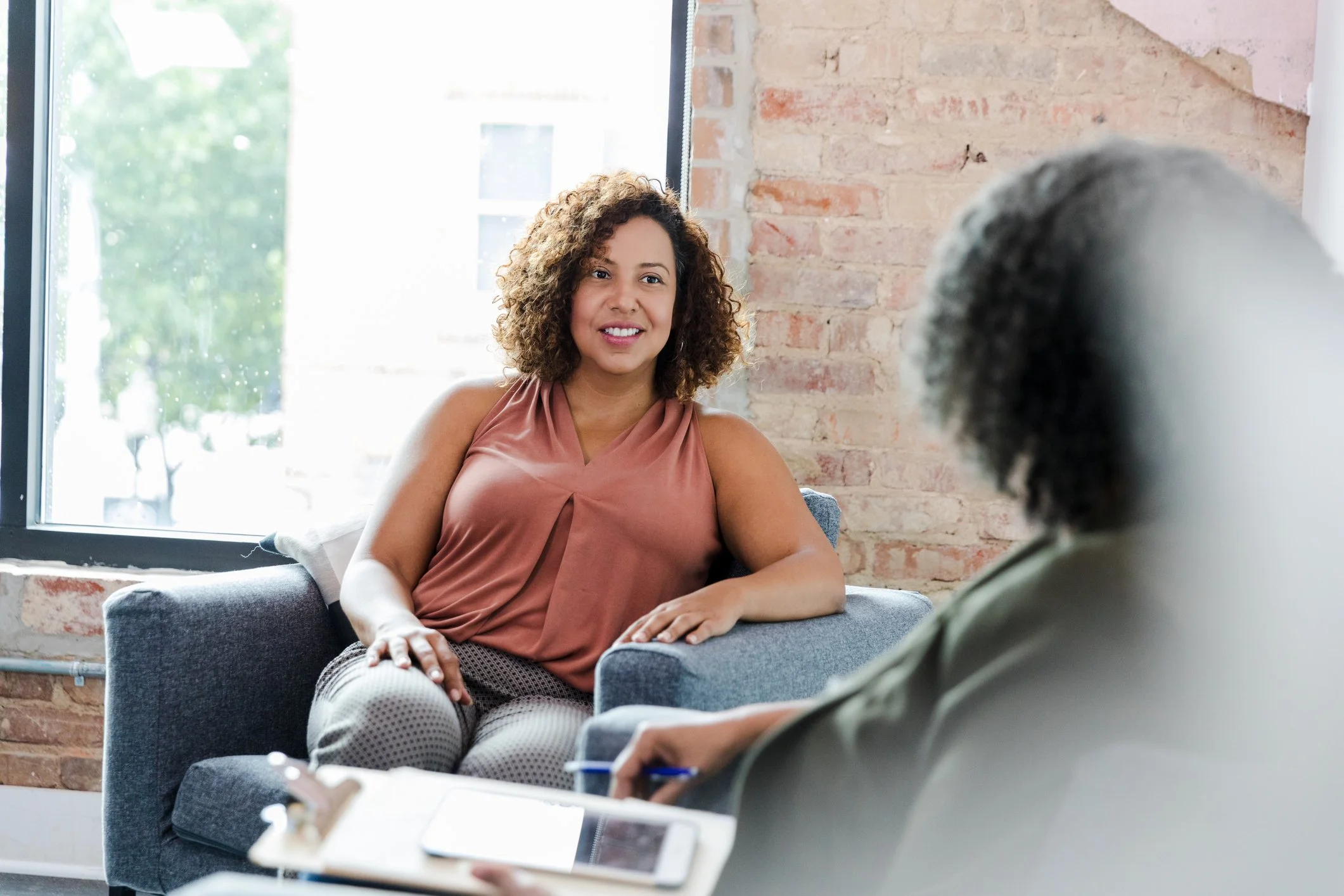A woman with curly hair in a sleeveless top is sitting on a sofa talking to another person, who is holding a phone. The setting appears to be a bright room with a brick wall and a large window.