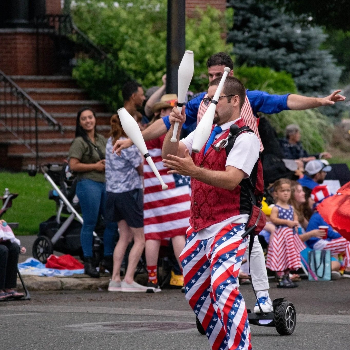 A man dressed in patriotic clothing juggling clubs during a celebration parade with people gathered along the sidewalk.