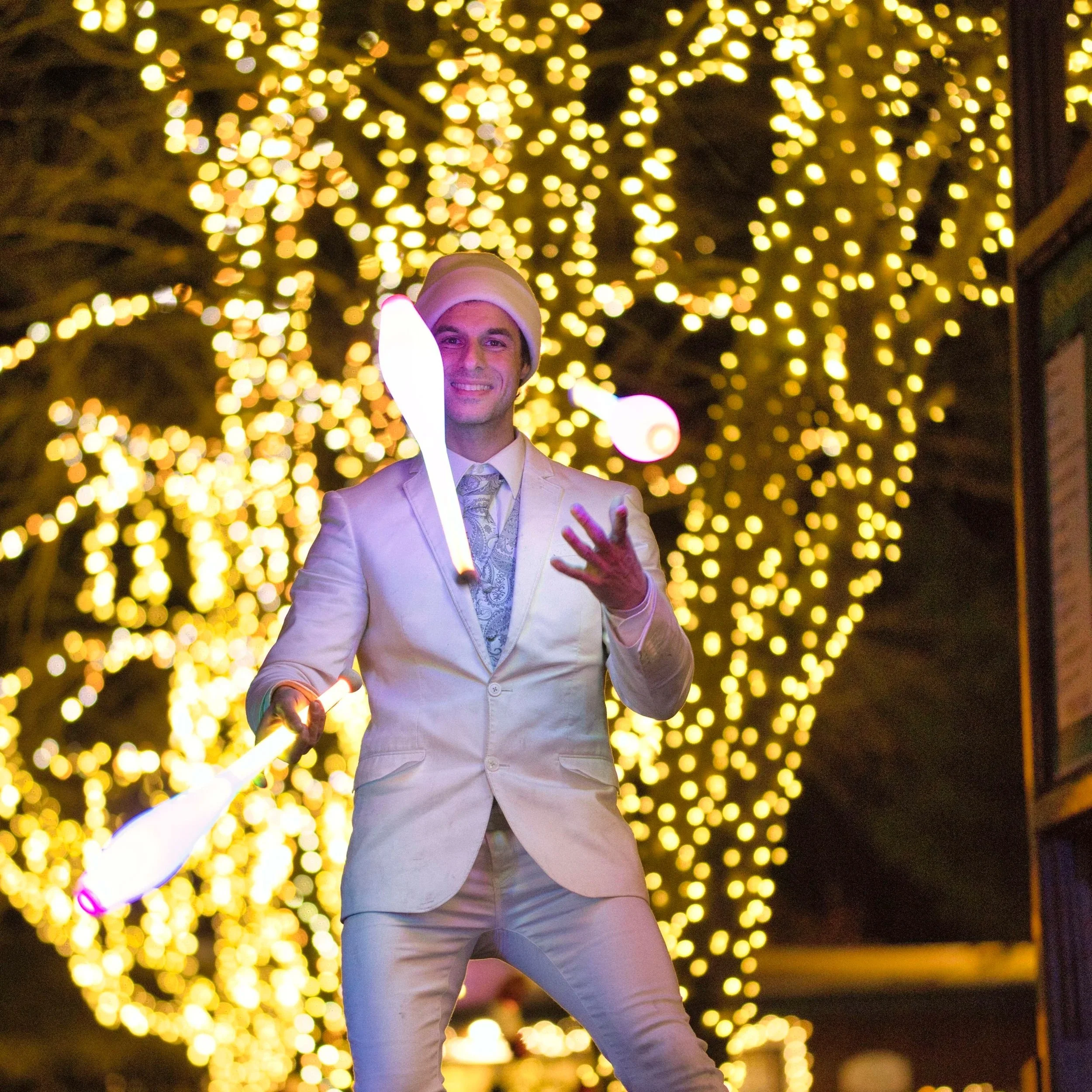 A man in a light suit and hat juggling glow sticks in front of a background of blurred Christmas lights.