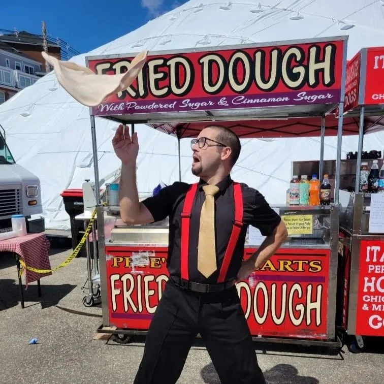 A man wearing glasses, a black shirt, red suspenders, and a tan tie is tossing a fried dough pastry in the air at a carnival stand. The stand has a red and black sign that reads 'Fried Dough' with powdered sugar and cinnamon sugar, and it's located outside.