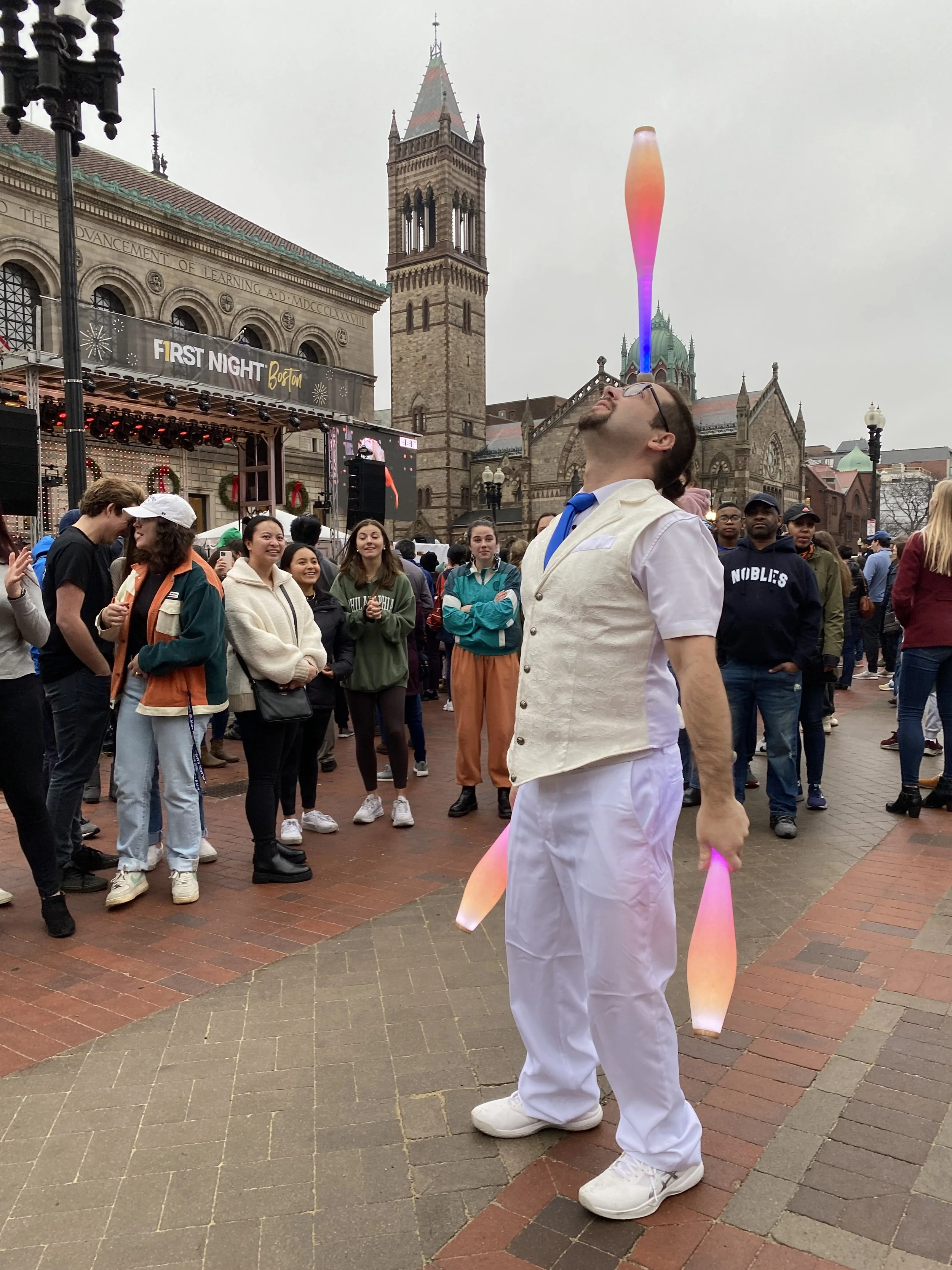 A man dressed as a juggler balances colorful lit juggling clubs on his face in a crowded outdoor area with historic buildings in the background.