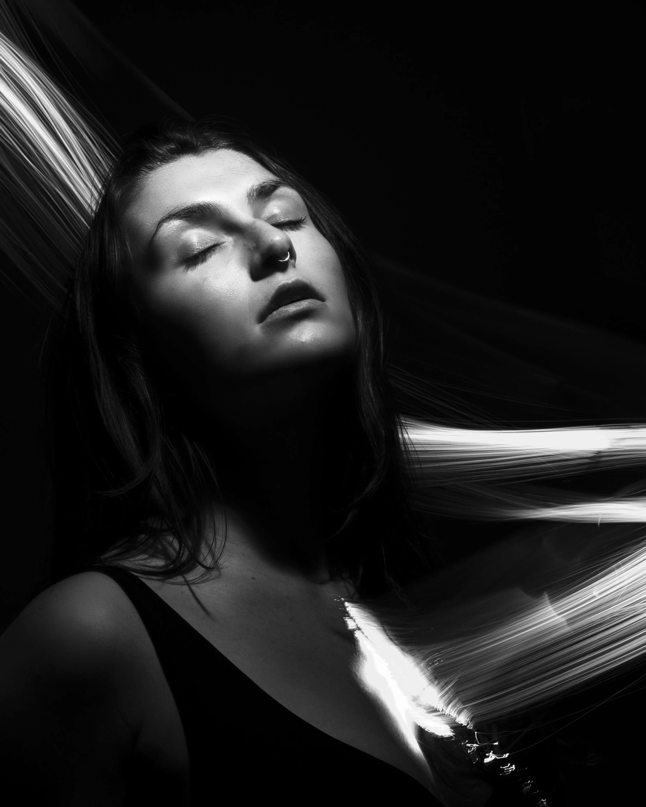 Black and white portrait of a woman with closed eyes and a necklace, with hair flowing and abstract streaks of light.