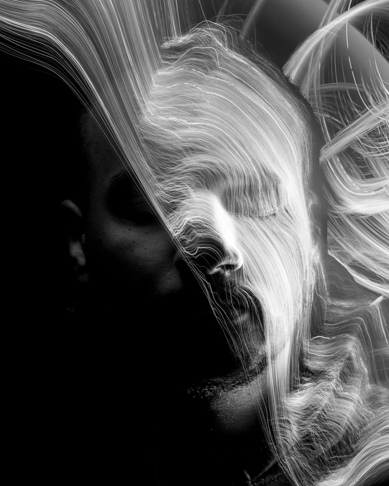 Black and white portrait of a person with long, flowing, light-colored hair, with some hair across their face, creating a flowing, artistic effect.